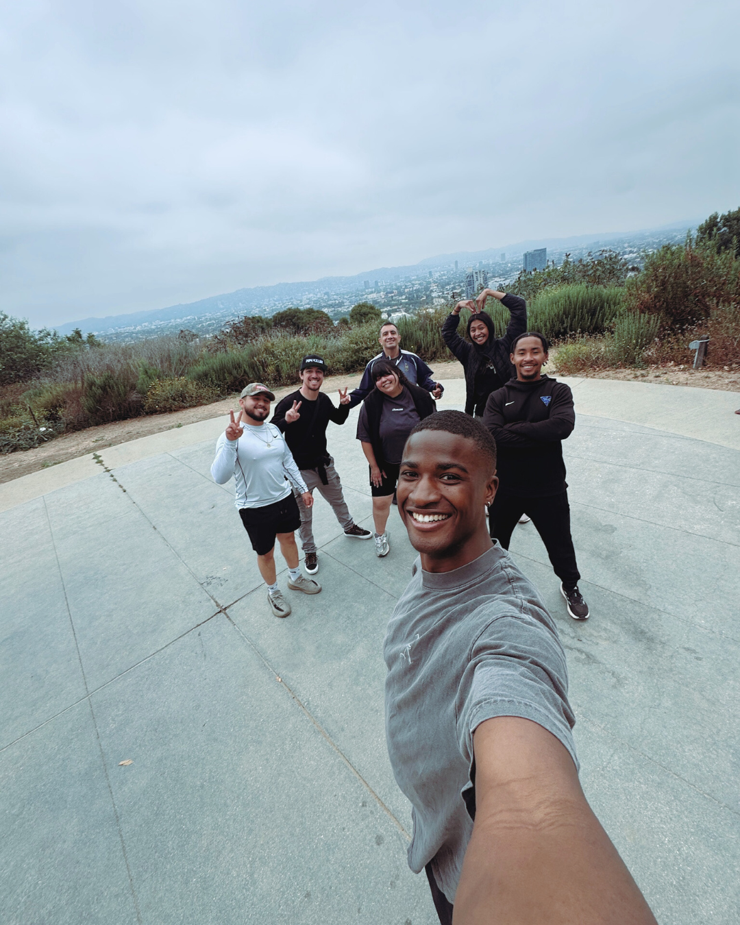 Group of seven friends taking a selfie on a hilltop with a cityscape and cloudy sky in the background.