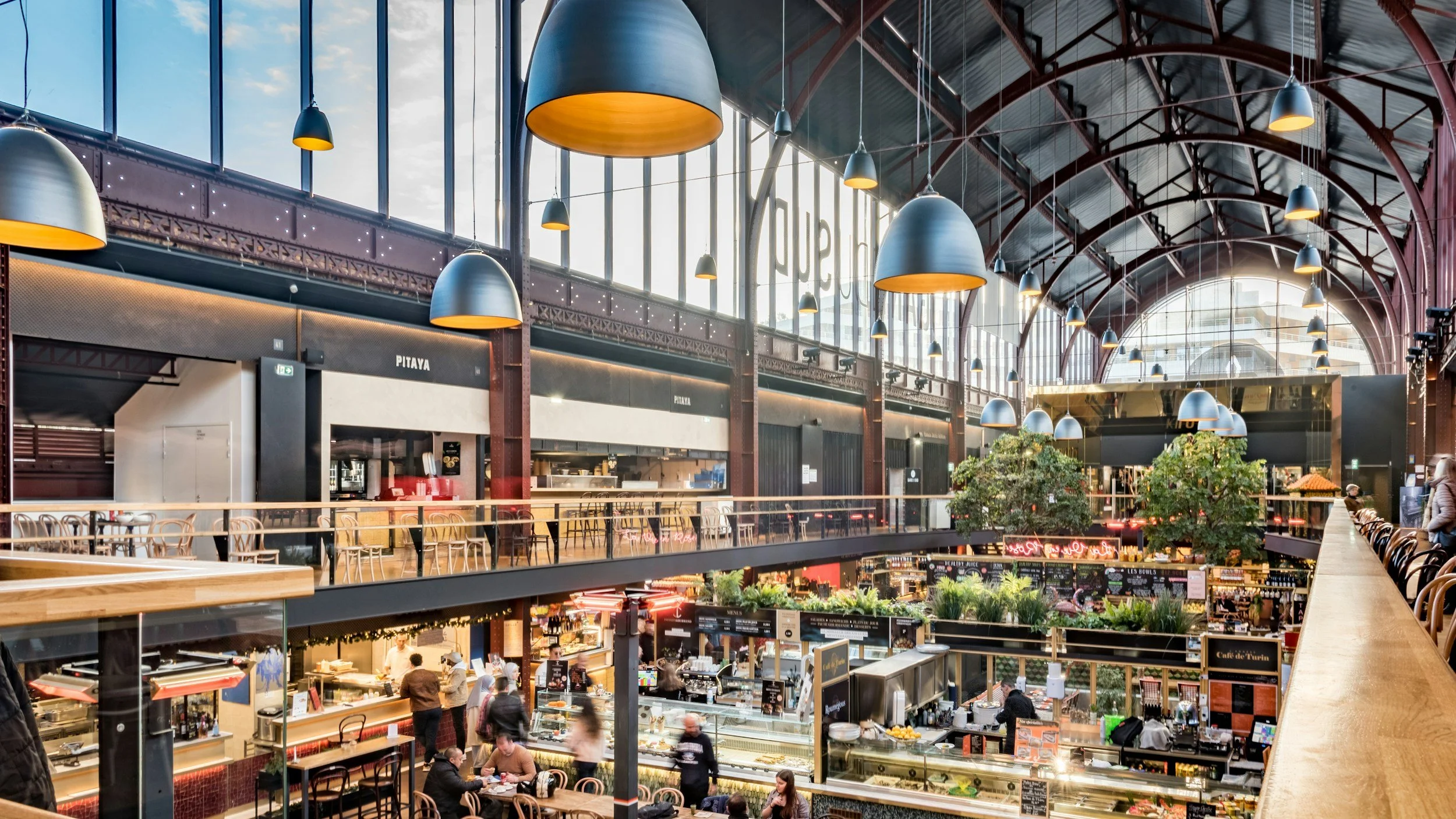A spacious indoor marketplace or food court with high arched glass ceiling, hanging pendant lights, and multiple food stalls with customers. Upper level has seating and trees for decoration.