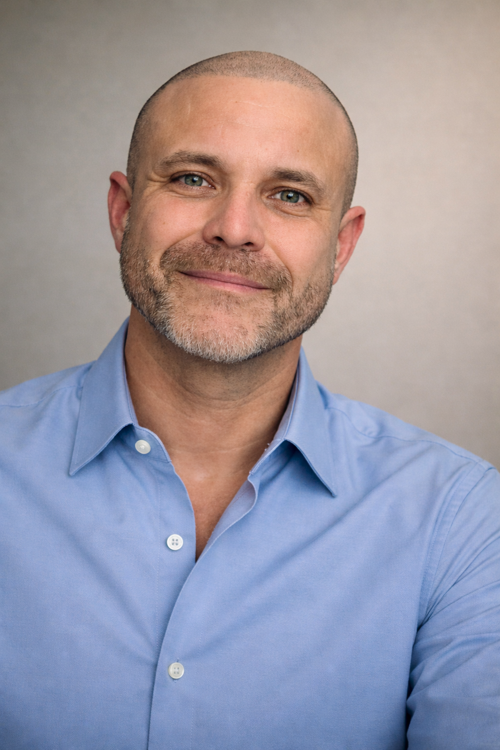 Close-up portrait of a middle-aged man with a shaved head and a beard, smiling and wearing a light blue button-up shirt, against a plain neutral background.