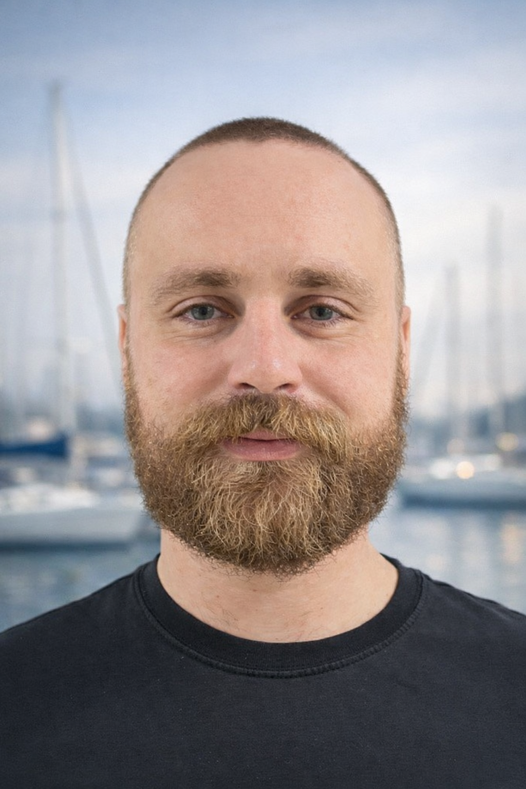 Portrait of a man with a beard and mustache, short hair, blue eyes, wearing a black shirt, with a marina in the background.