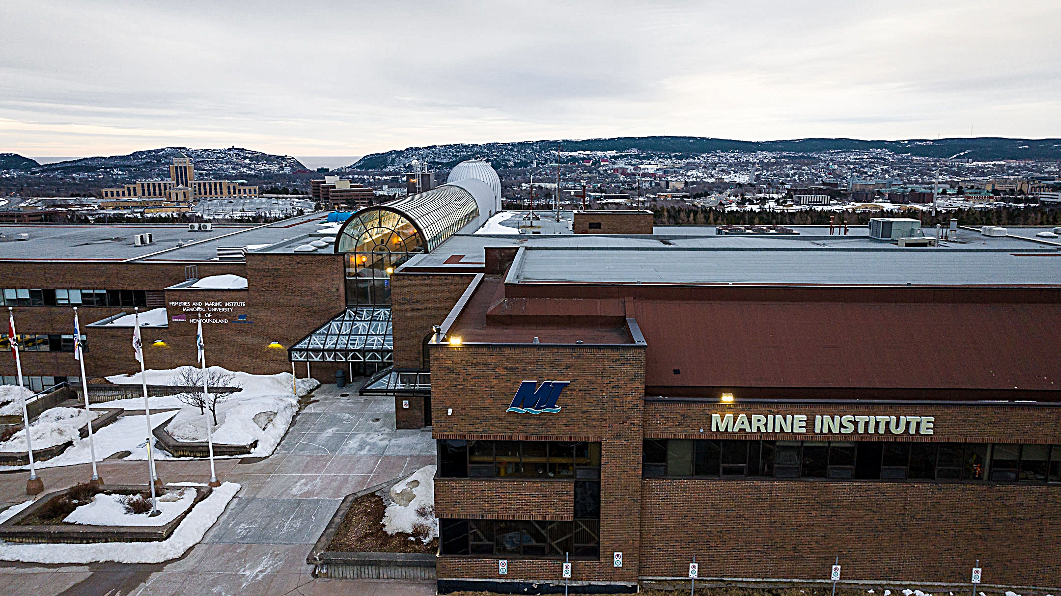 Aerial view of the Fisheries and Marine Institute at Memorial University of Newfoundland during winter, showing snow-covered grounds, flags, brick building with signage, and a distant cityscape with hills.