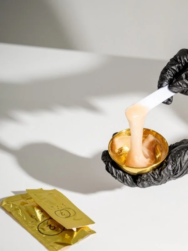 Person wearing black gloves holding a spatula and stirring pink wax in a small gold container with two gold-colored packets nearby on a white surface.