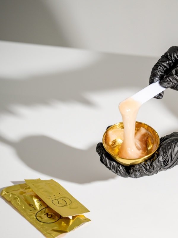 Person wearing black gloves holding a small golden container with a spatula scraping a light pink or peach-colored substance, with gold-colored packaging on a white surface.