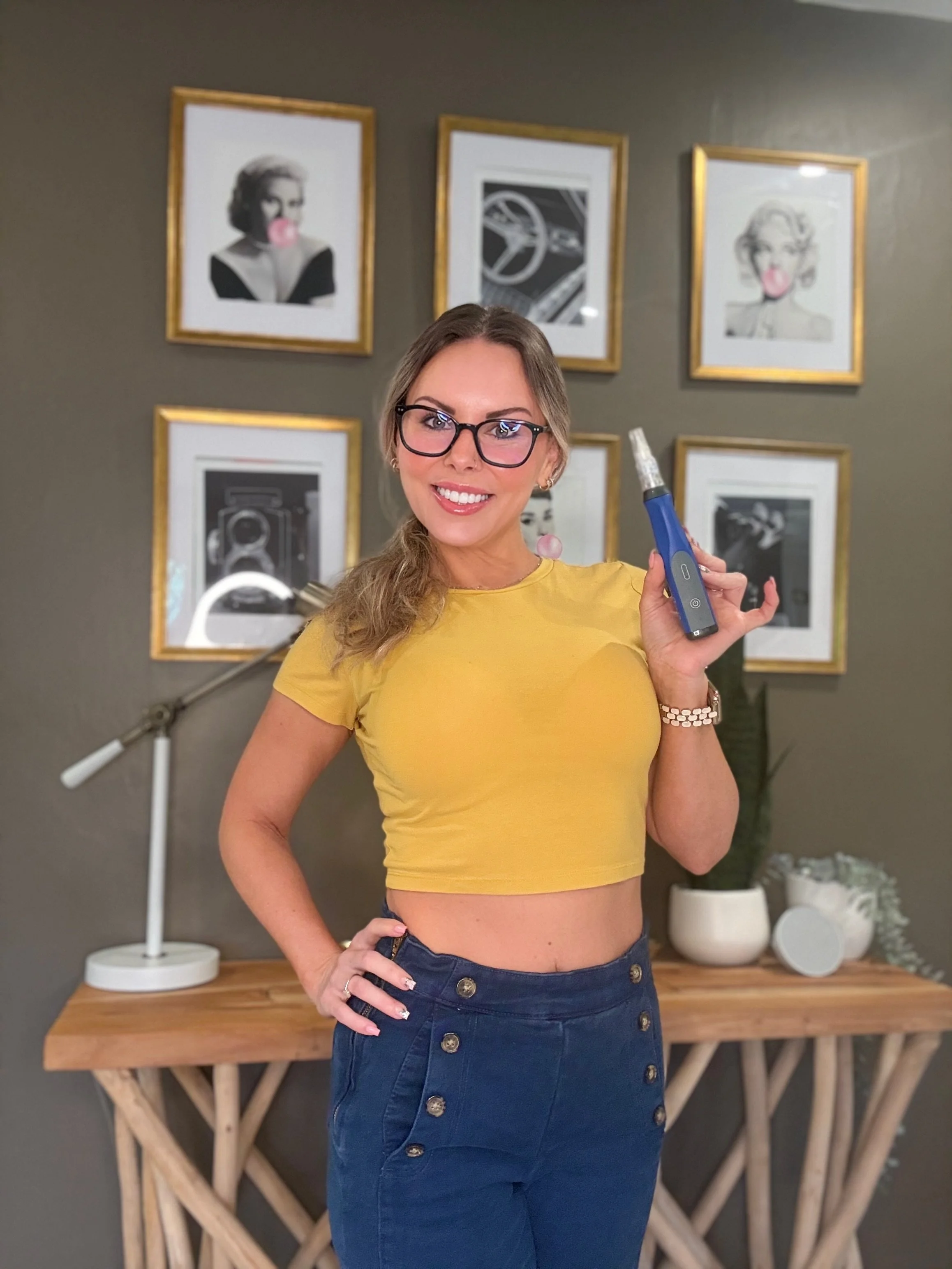 A woman with glasses wearing a yellow crop top and navy blue skirt stands in a room with a wooden table and framed pictures on the wall behind her, holding a black and blue electric soldering iron.