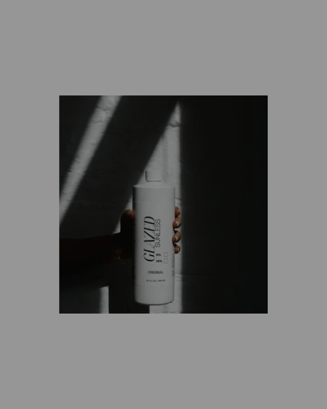 A woman with long brown hair wearing earrings and a watch, standing outdoors with sunlight, holding her hair back with one hand, and wearing a light-colored top against a beige wall and blue sky.