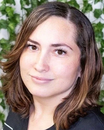 A woman with shoulder-length brown hair and light skin, smiling, standing in front of green foliage.