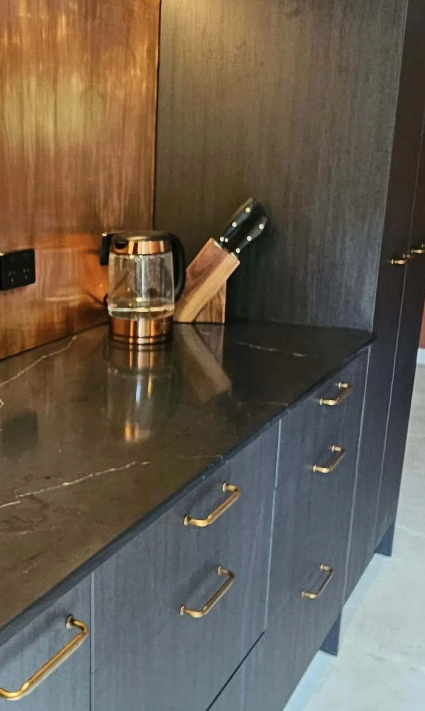 A kitchen counter with a glass teapot, a knife block, and dark cabinets with gold handles.