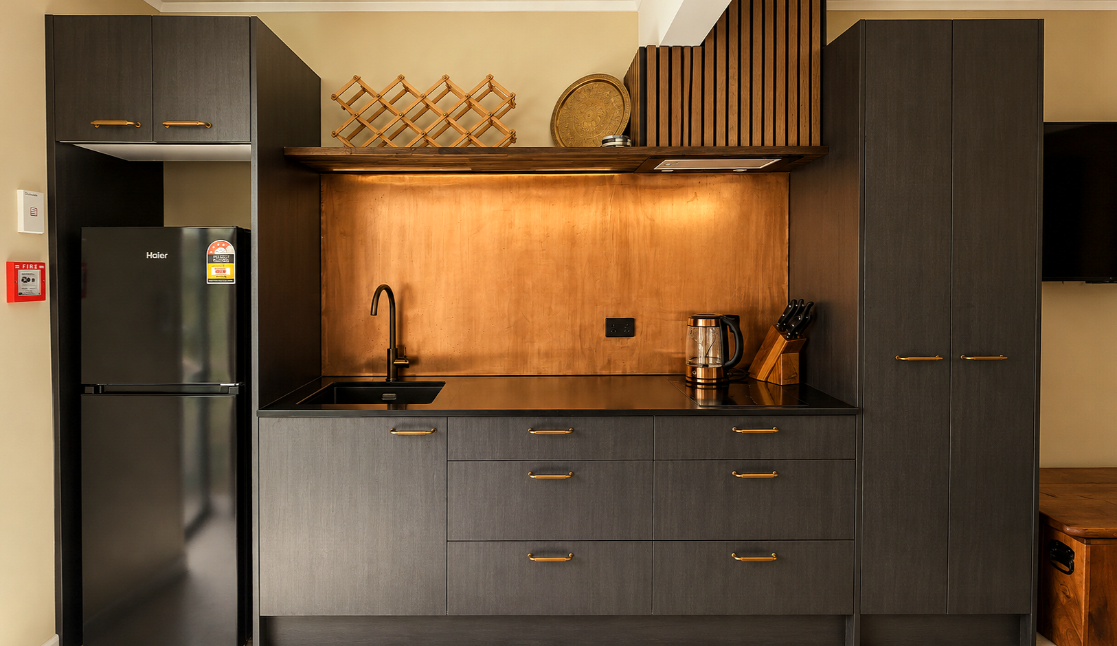 Modern kitchen with black cabinets, wooden backsplash, stainless steel sink, black faucet, and black refrigerator. Items on the counter include a clear electric kettle and a wooden knife block. Open shelves above display wooden wine racks and a decorative tray.