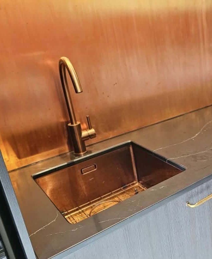 A kitchen sink with a brass faucet and a copper-colored interior, set in a black marble countertop with a wooden backsplash.