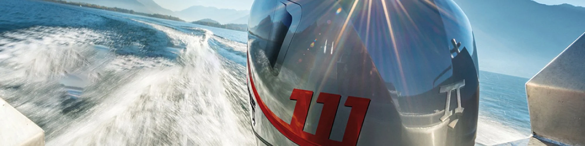 View of a boat's outboard motor with the number 111, moving through the water on a sunny day with a clear sky and distant shoreline.