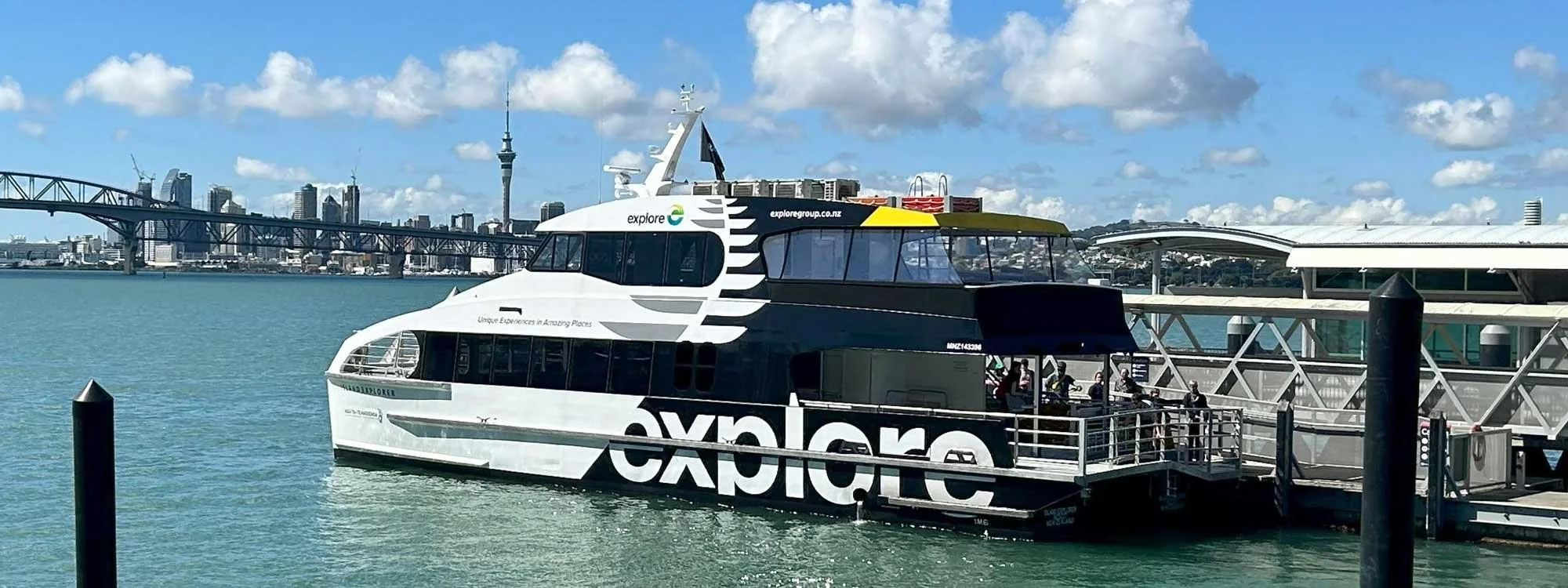 A white and black Explore ferry boat docked at a pier with city skyline and bridge in the background, under a partly cloudy sky.
