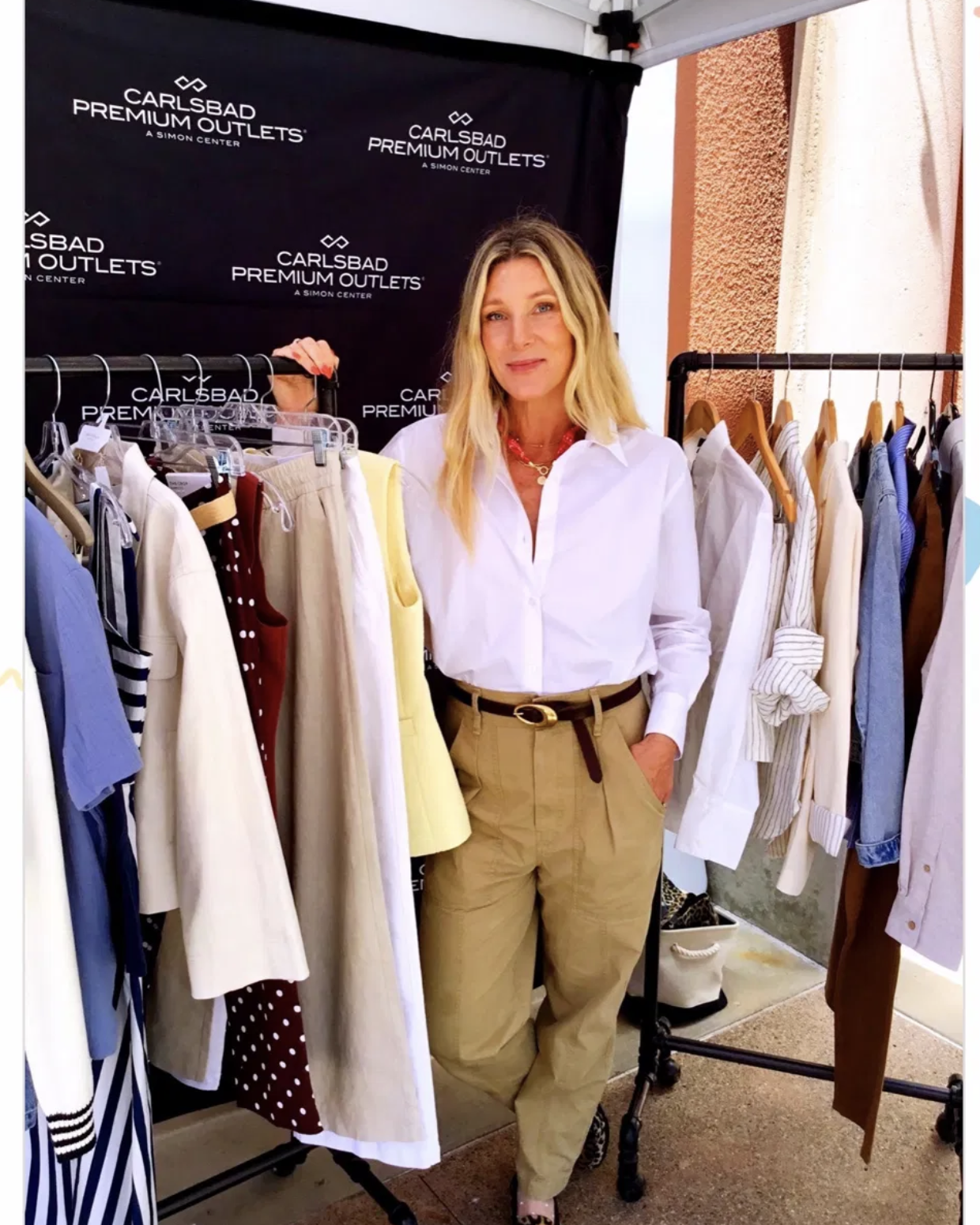 A woman standing between two clothing racks filled with various women's clothing items, smiling at the camera, at Carlsbad Premium Outlets.