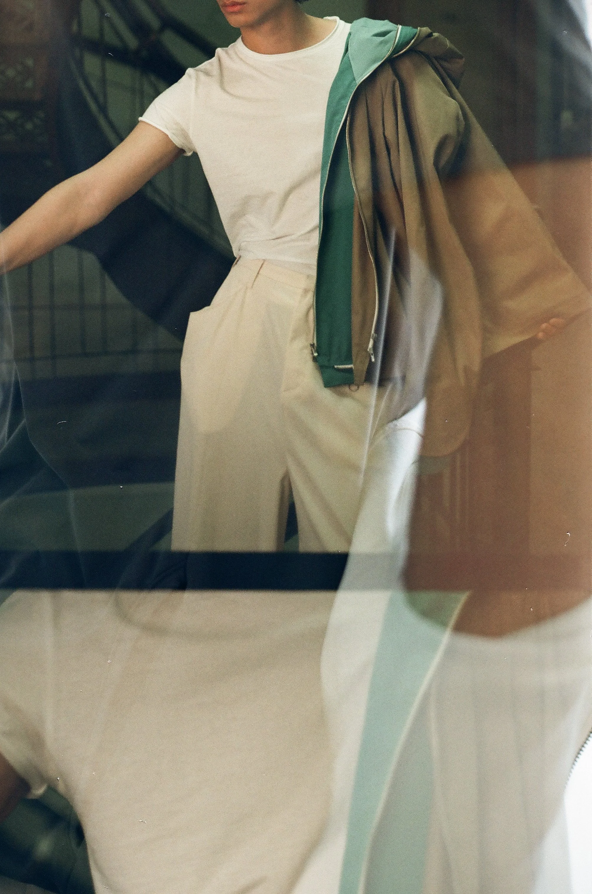 Person wearing a white t-shirt and beige pants, holding a green jacket over their shoulder, seen through a glass window with reflections.