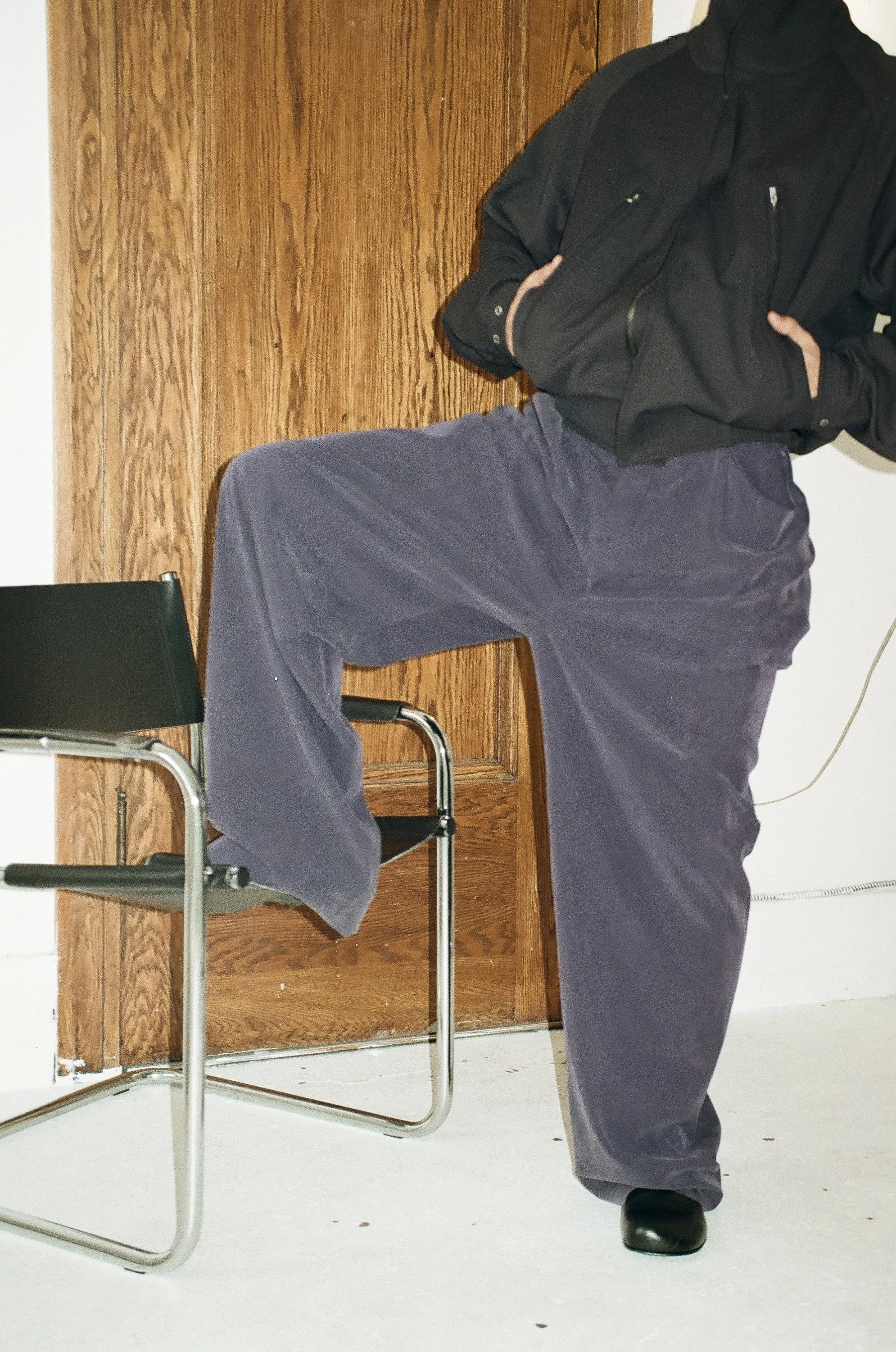 Person standing with one foot on a black chair, wearing a black jacket and loose dark pants, in a room with a wooden door and white walls.