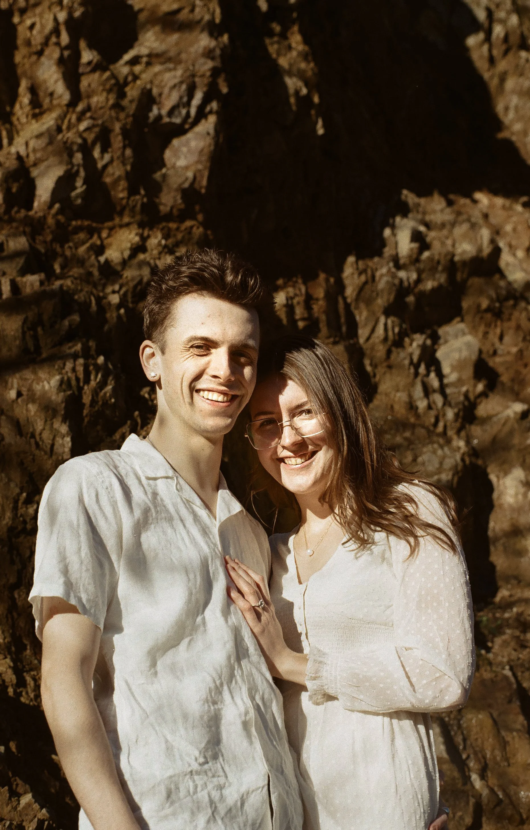 A smiling young man and woman standing close together outdoors at sunset with rocky background.