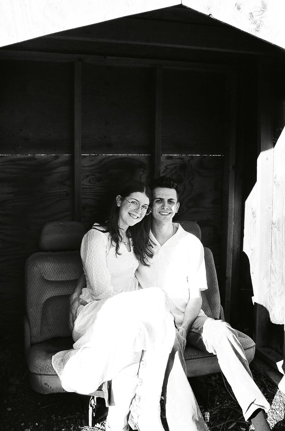 A black-and-white photo of a smiling couple sitting close together on a cushioned bench inside a small wooden structure, possibly a playhouse or garden shed, with simple wooden walls and a peaked roof.