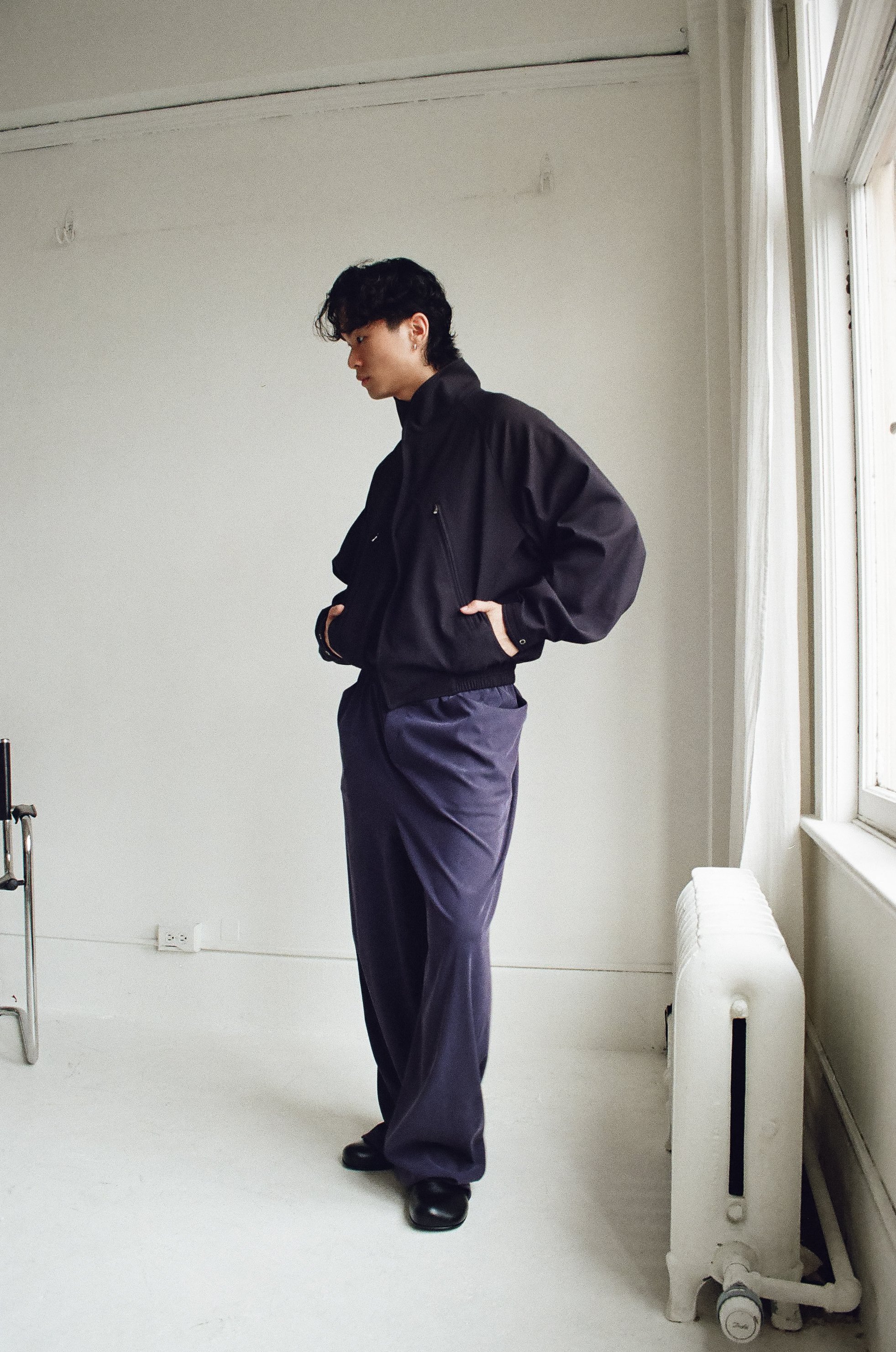 Young man with dark, curly hair in a black jacket and baggy purple pants stands indoors near a window, with his hands in his jacket pockets, looking down.