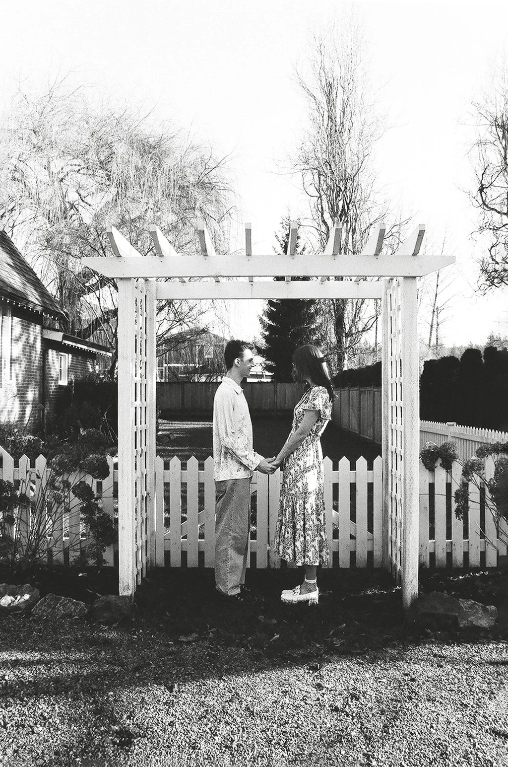 A black and white photo of a couple holding hands and facing each other under a white wooden arbor in a garden. The woman wears a long dress and the man wears a light shirt and pants. A white picket fence surrounds the garden, with trees in the backg