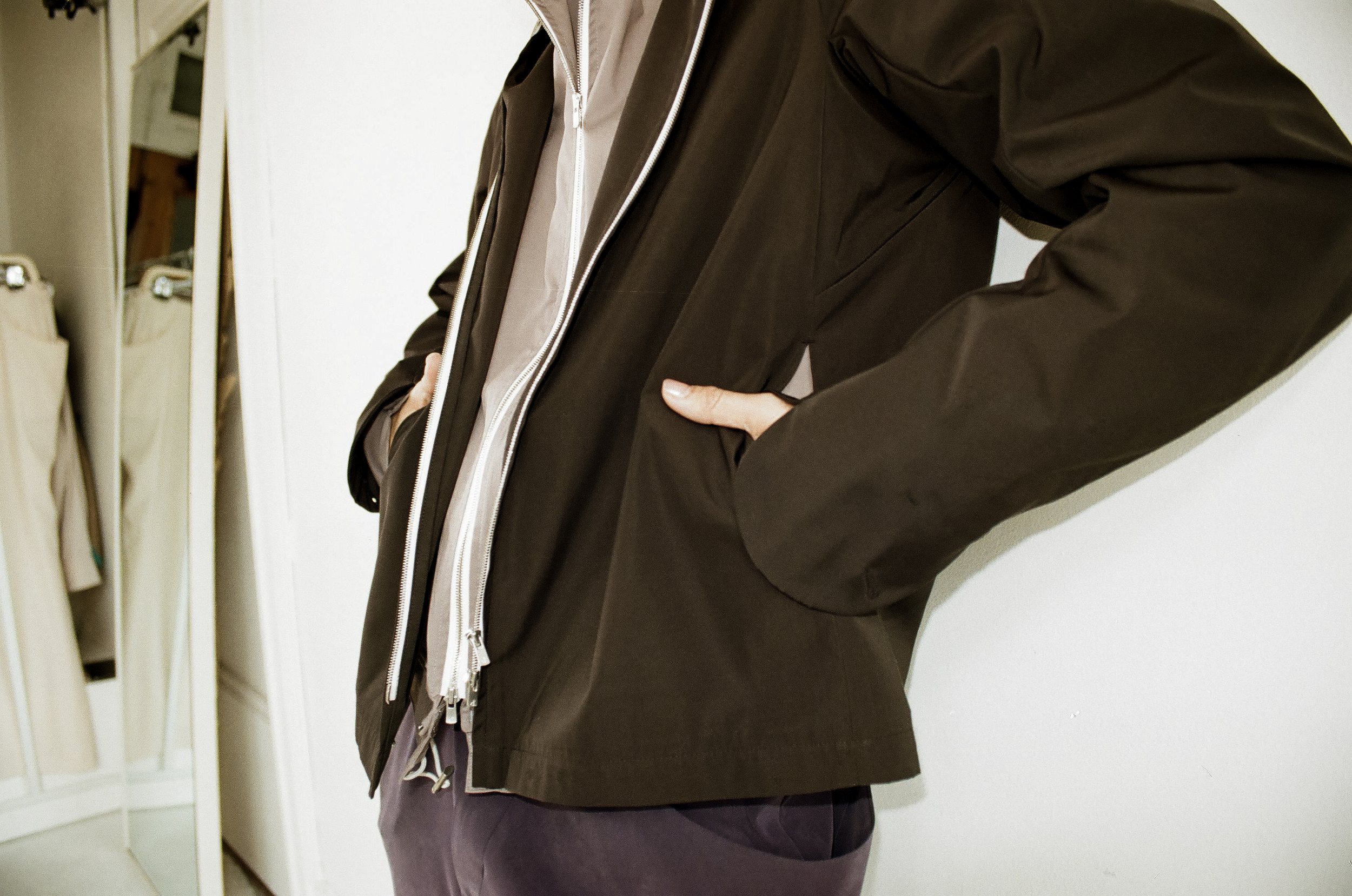 Person wearing a dark jacket with hands in pockets standing against a white wall.