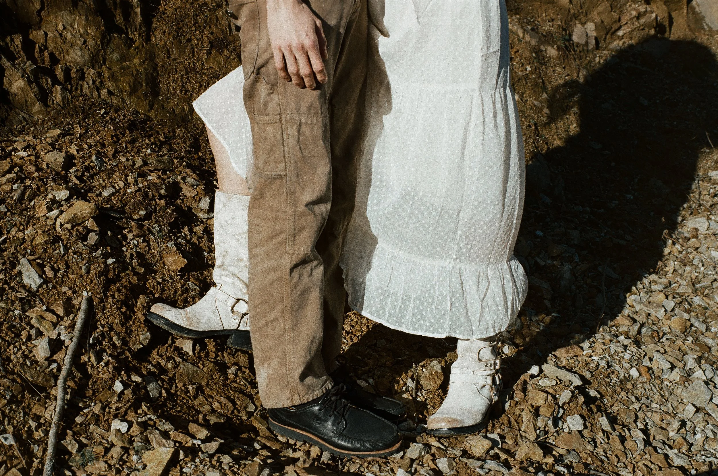 Close-up of a couple standing on rocky ground, focusing on their lower bodies. The woman is wearing white boots and a white dress, while the man is wearing beige pants and black shoes.