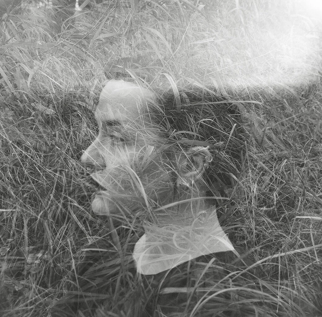 A double exposure black and white photo of a woman lying in tall grass, with her profile face visible.