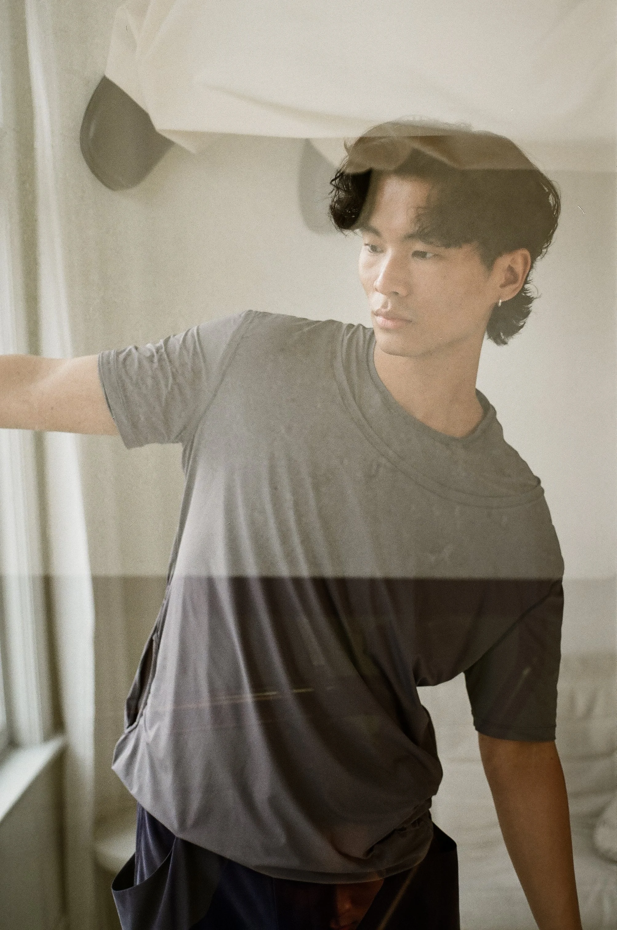 A young man with dark hair and an earring wearing a grey shirt, standing indoors near a window, partially obscured by a reflection or glass.