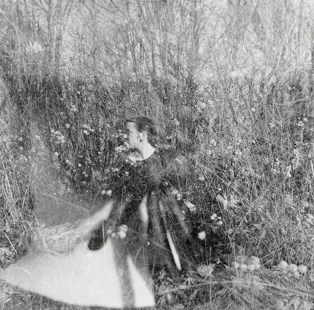 A woman with dark hair standing among tall, dense, dried grass and plants in a natural outdoor setting.