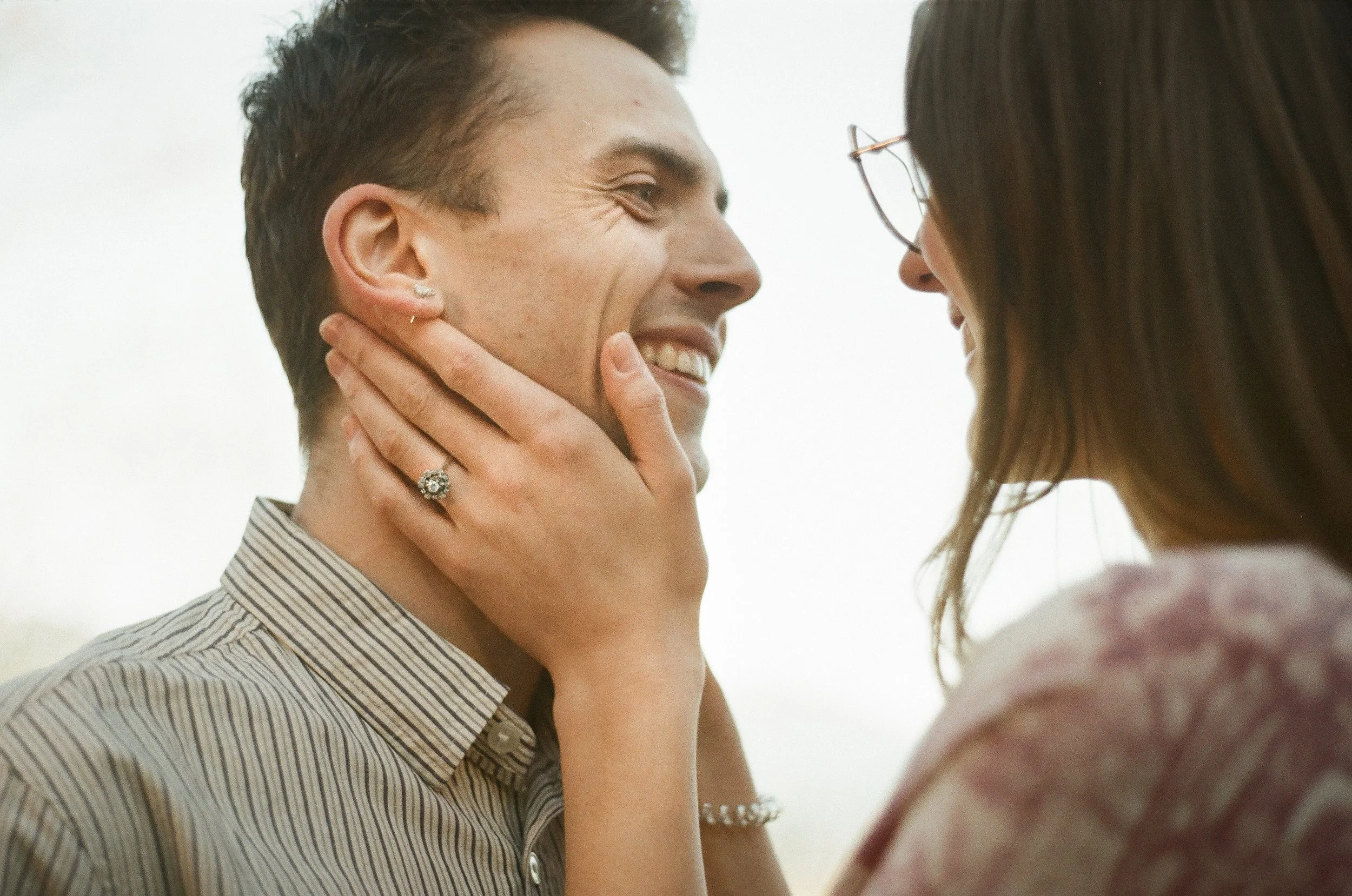 A woman holding a man's face gently with both hands, smiling and making eye contact outside.