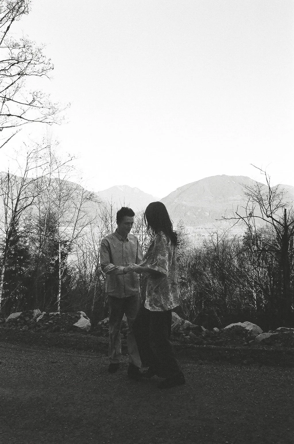 A black and white photo of a couple standing outdoors with a mountain and trees in the background. The couple is holding hands and facing each other.