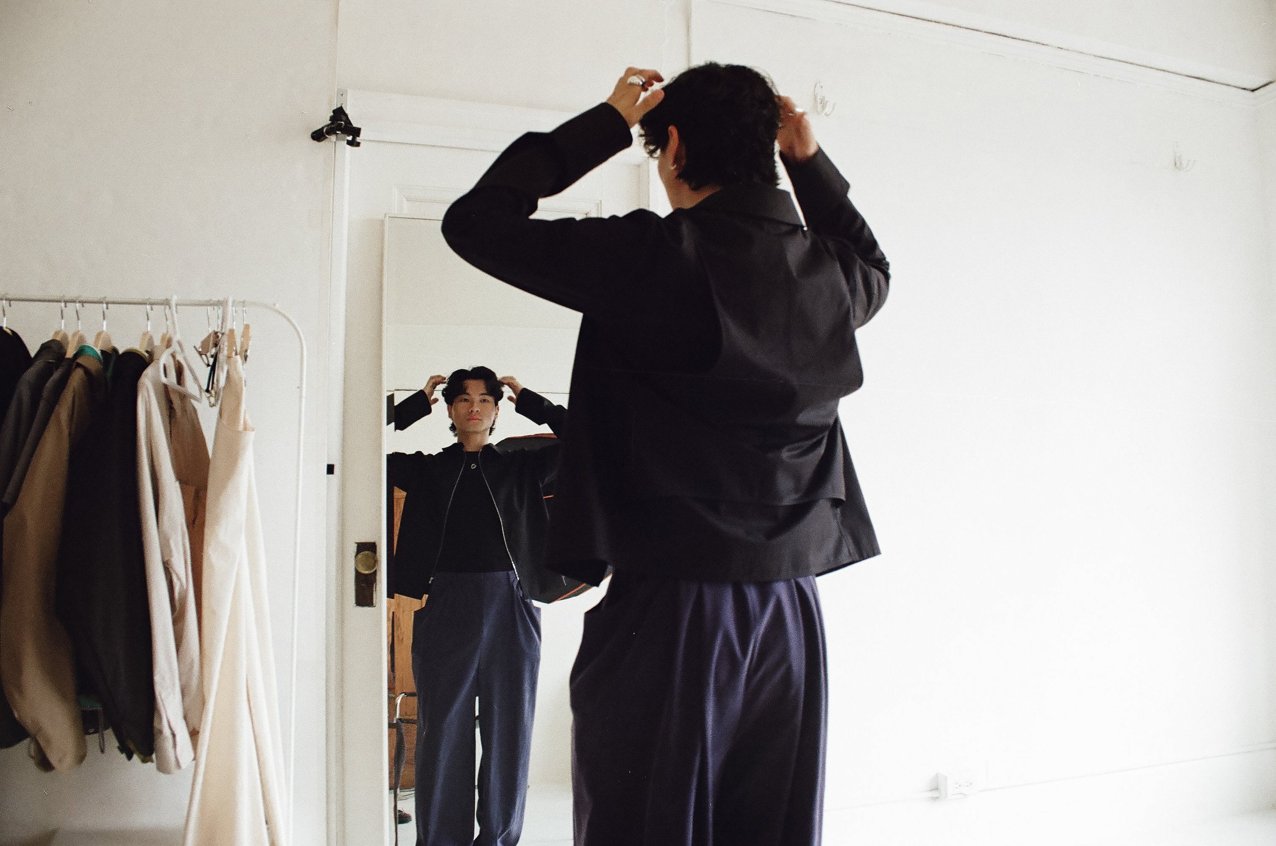 Person with curly hair looking in a mirror, adjusting their hair, standing in a room with beige and white walls, clothing rack with jackets on the side.
