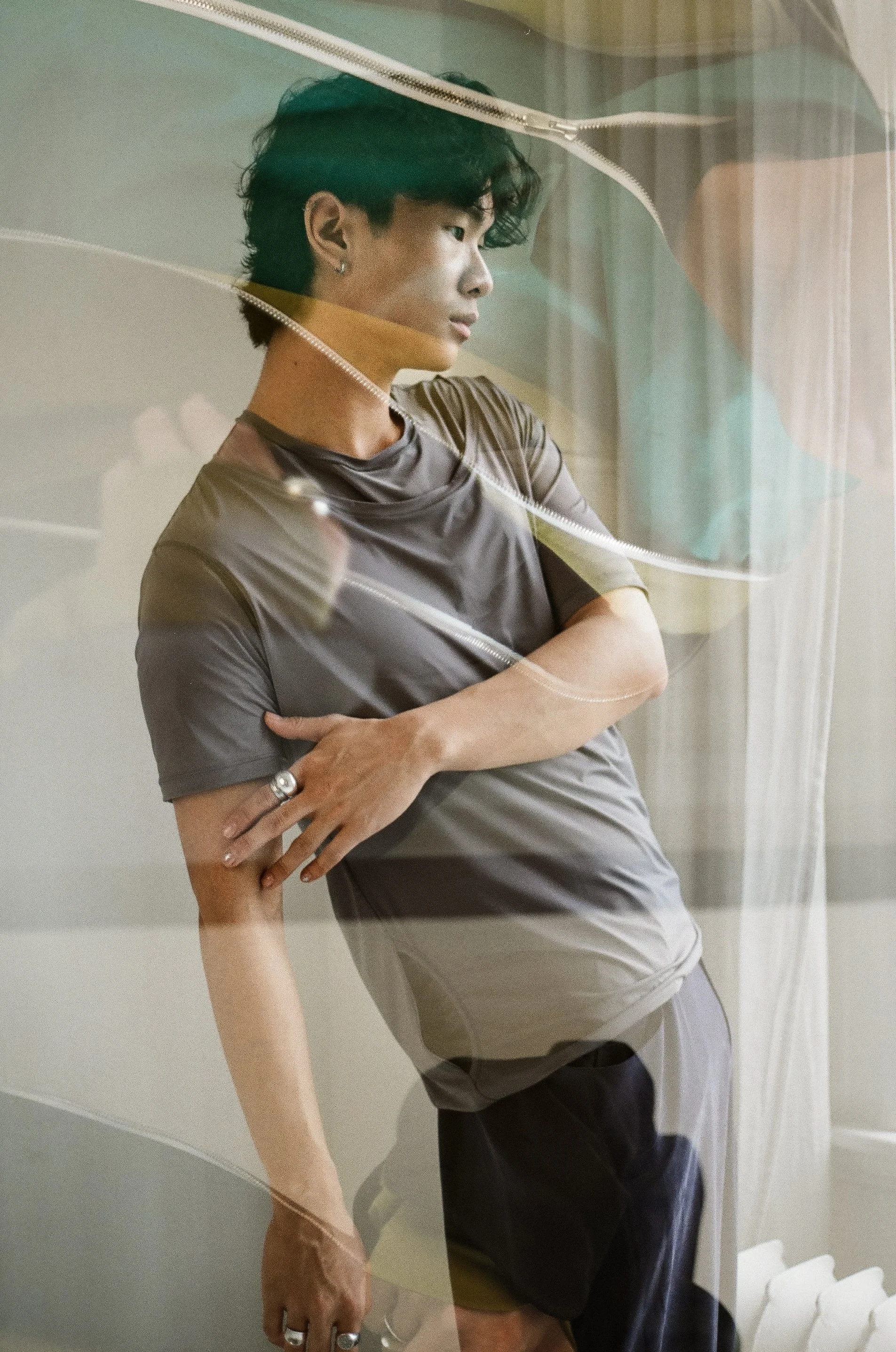 A young person with dark, wavy hair and earrings leaning against a glass window, with a contemplative expression.