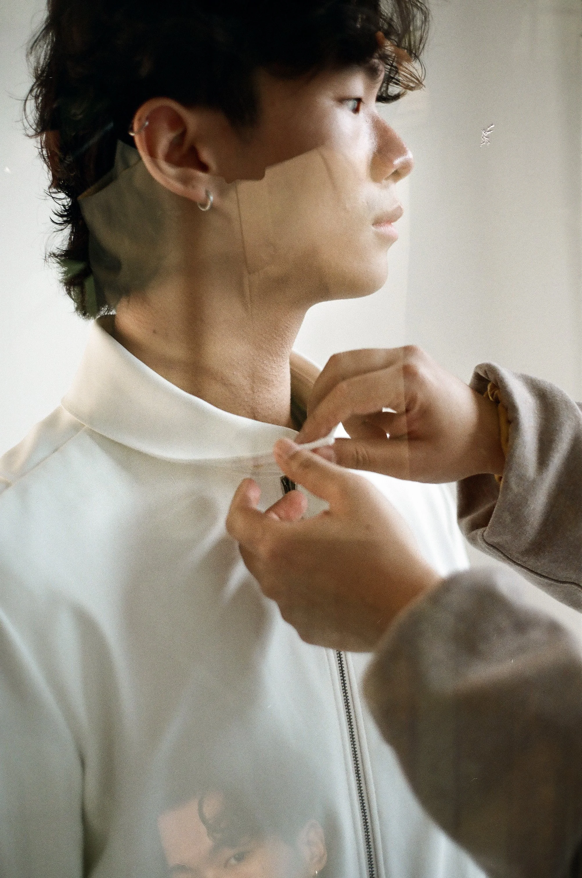 A young man with dark, wavy hair is having the collar of his light-colored jacket zipped up by another person. He is facing to the right, and a small part of his face and ear are visible. The scene appears to be indoors with neutral lighting.