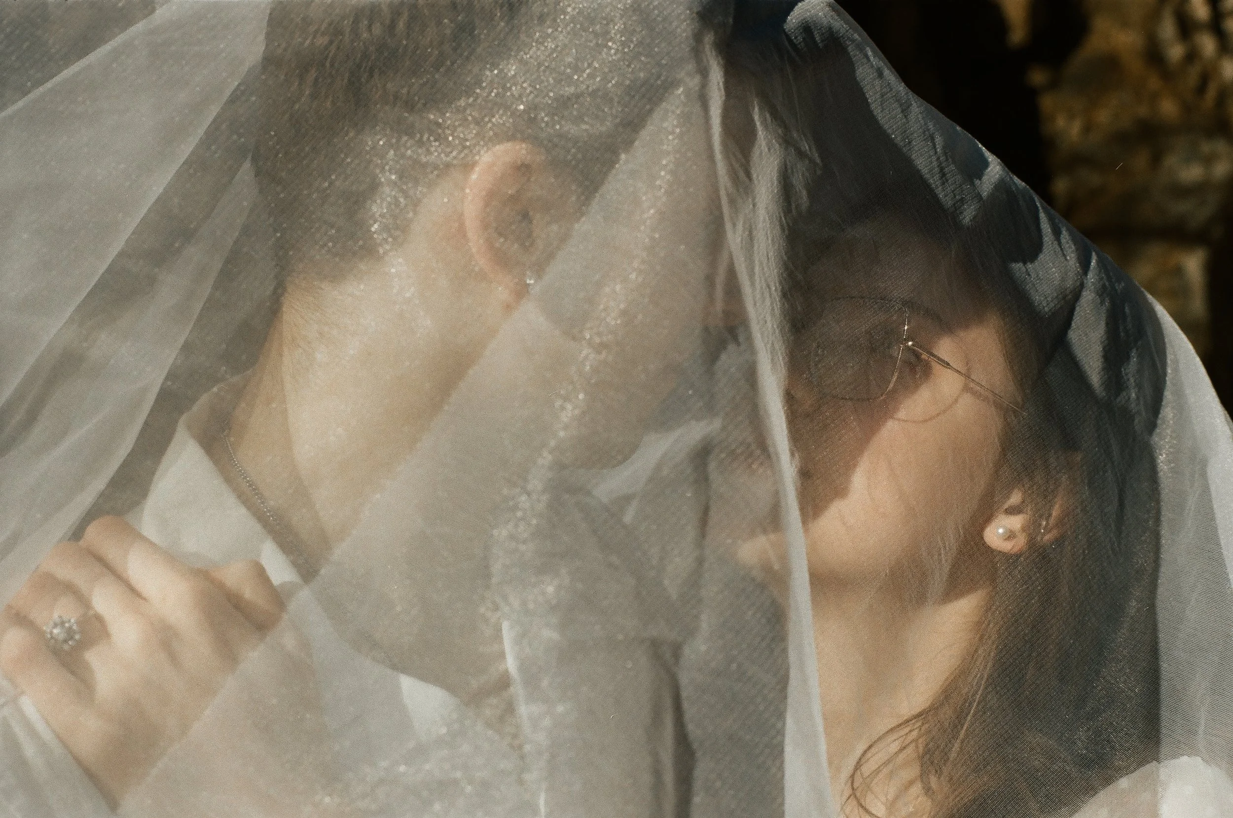 A couple faces close under a sheer veil, with one person's hand visible wearing a ring, both wearing glasses and having dark hair.
