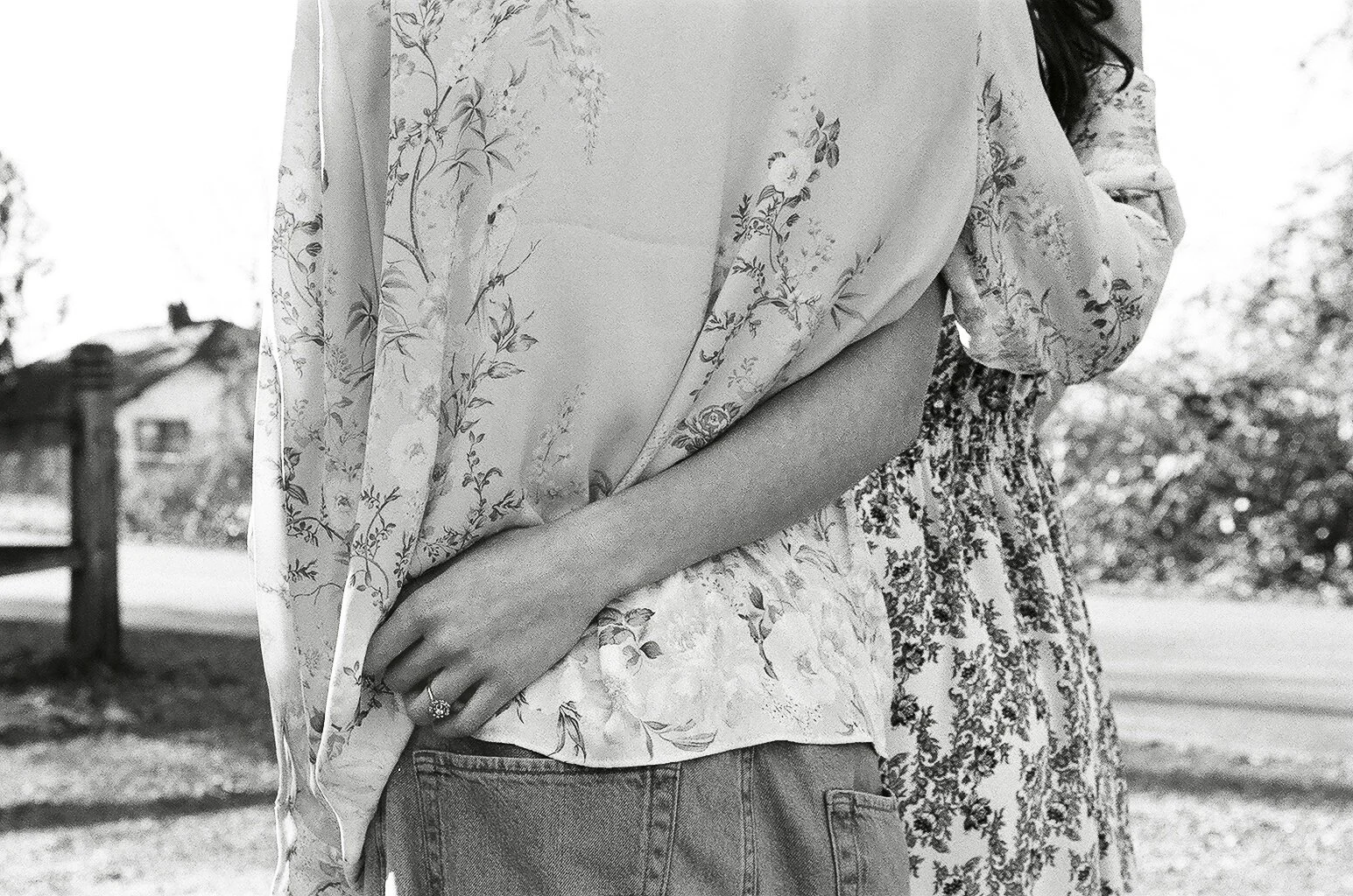 A black-and-white photo of two women standing outdoors, one in the foreground and the other slightly behind. The woman in the foreground is wearing a floral blouse, displaying her left hand with a ring. The woman behind is wearing a floral dress. Tre