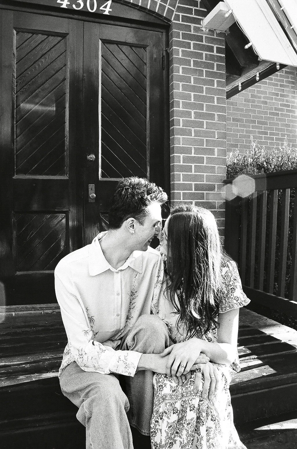 A black and white photo of a man and a woman sitting on a bench outside a brick building, leaning close and touching hands, with their foreheads and noses almost touching, sharing an intimate moment.