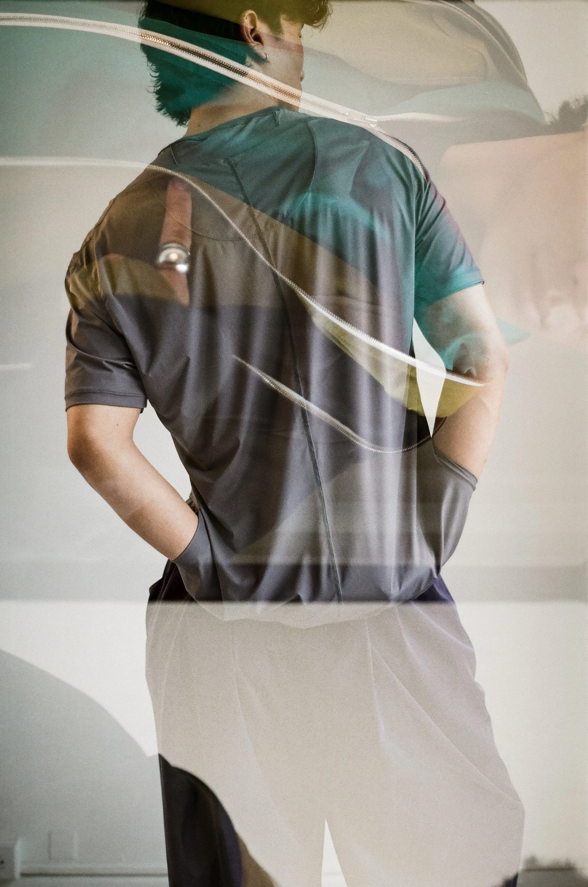 A person wearing a black shirt and gray pants with hands in pockets, seen from the back through a glass window or mirror.