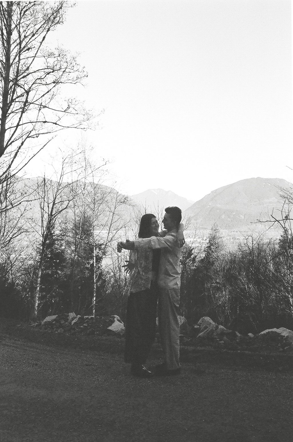 A couple hugging in a mountain landscape with trees and mountains in the background, black and white photograph.