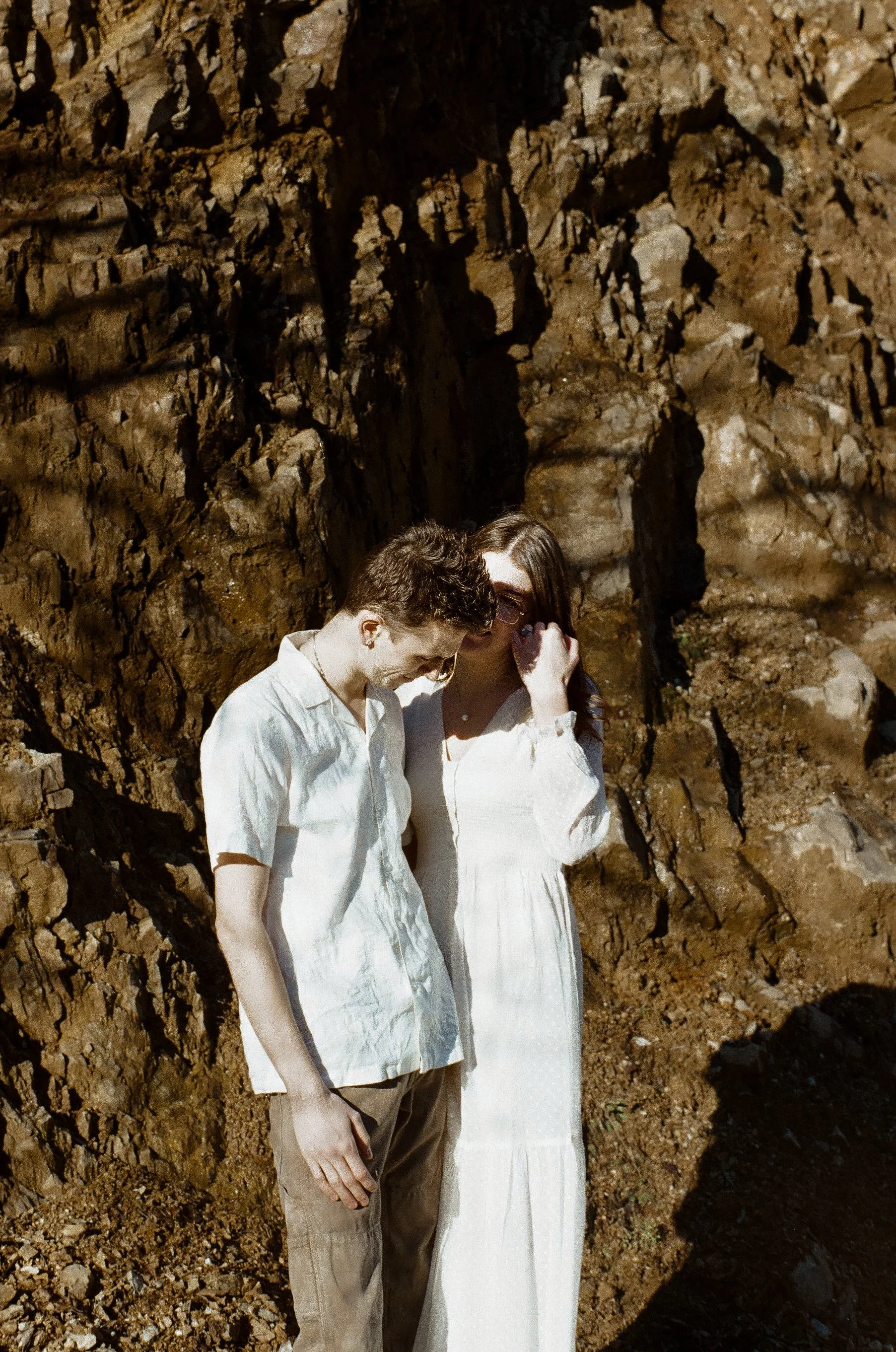 A young couple in light-colored clothing standing close together outdoors against a rocky background.