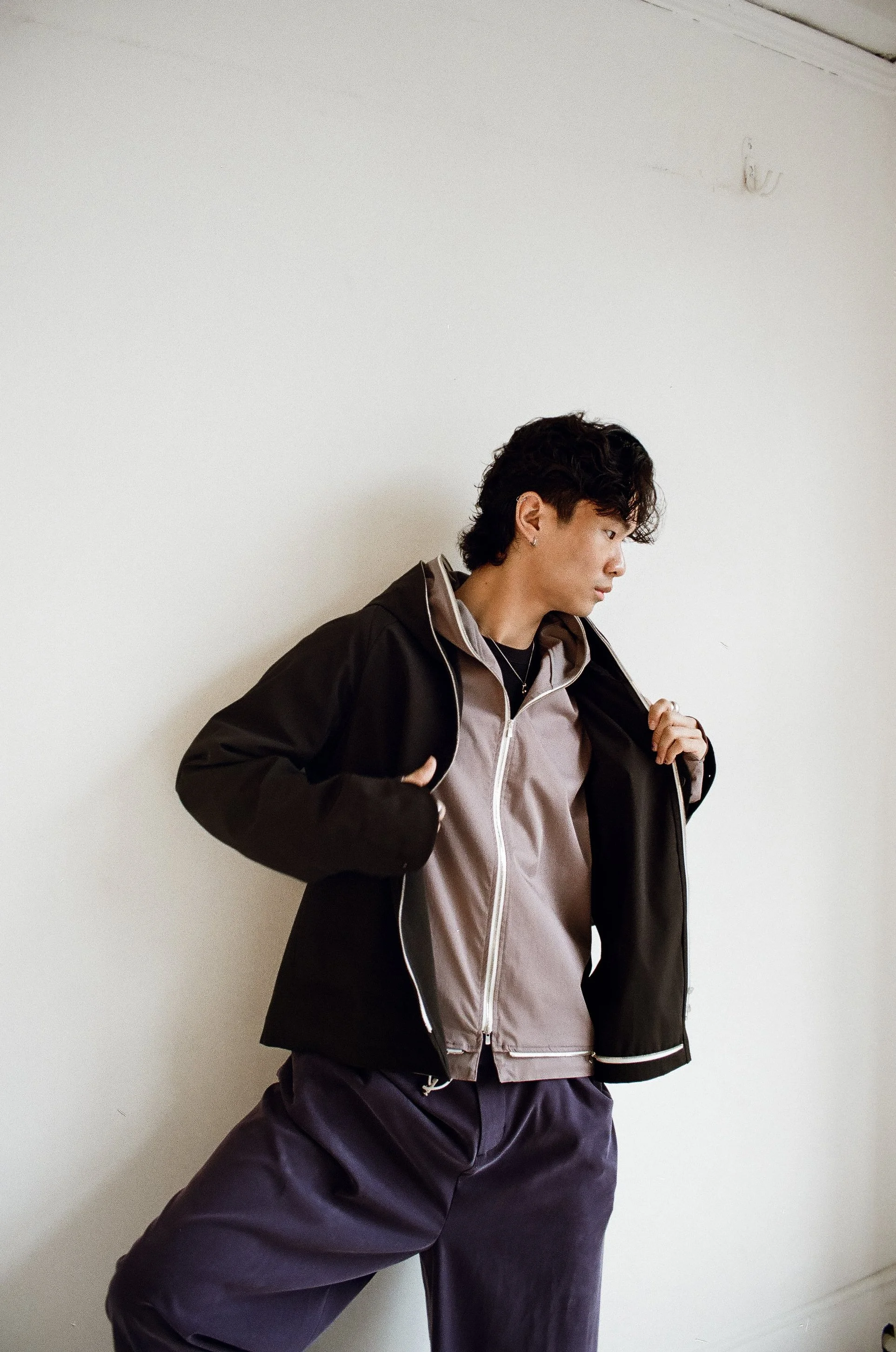 A young man with dark, curly hair wearing a black and gray jacket over a gray zip-up hoodie, standing against a plain white wall, looking to the side with one hand on his jacket.