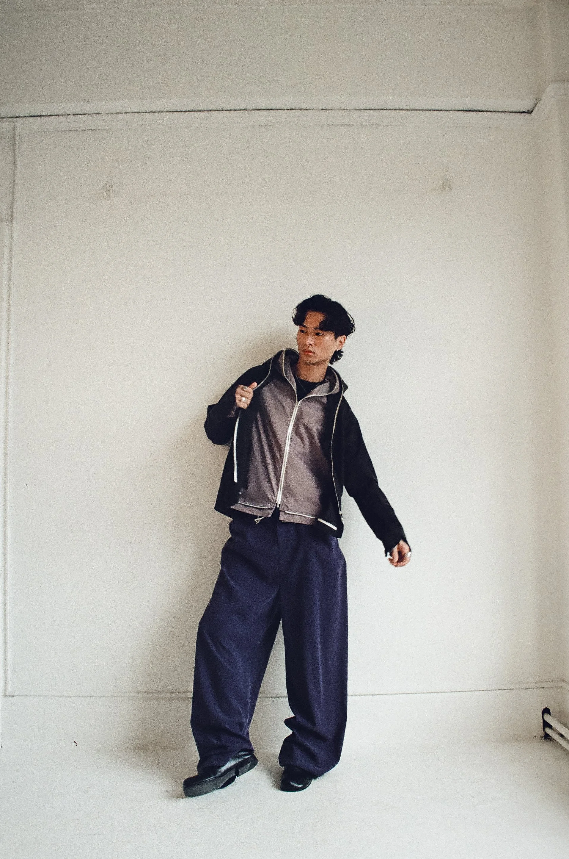 A young man standing against a white wall, wearing a black and gray jacket, dark wide-leg pants, and black shoes, holding a backpack over his shoulder.