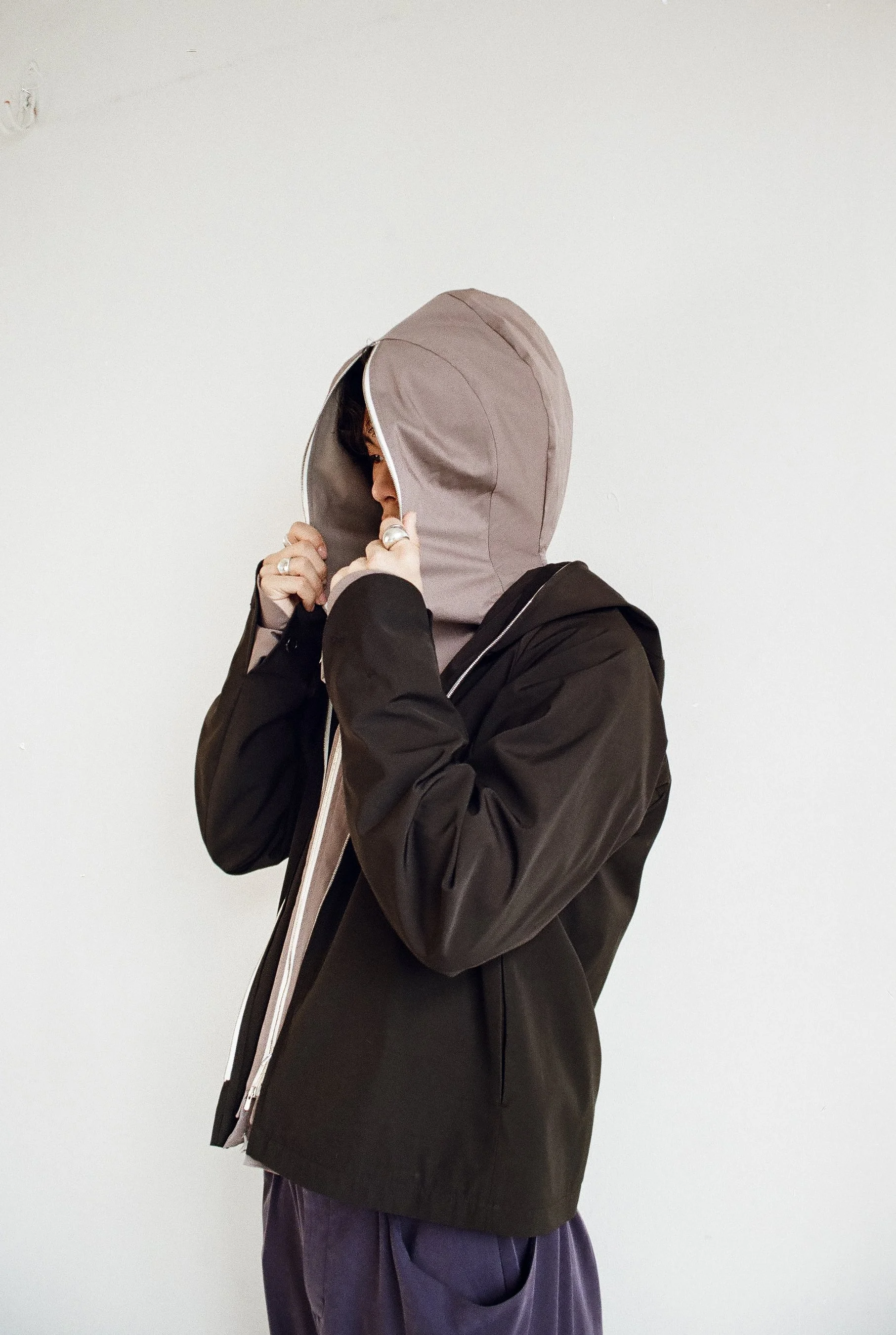 A person wearing a black jacket with a beige hood, standing against a plain white wall, holding the hood closed with both hands.