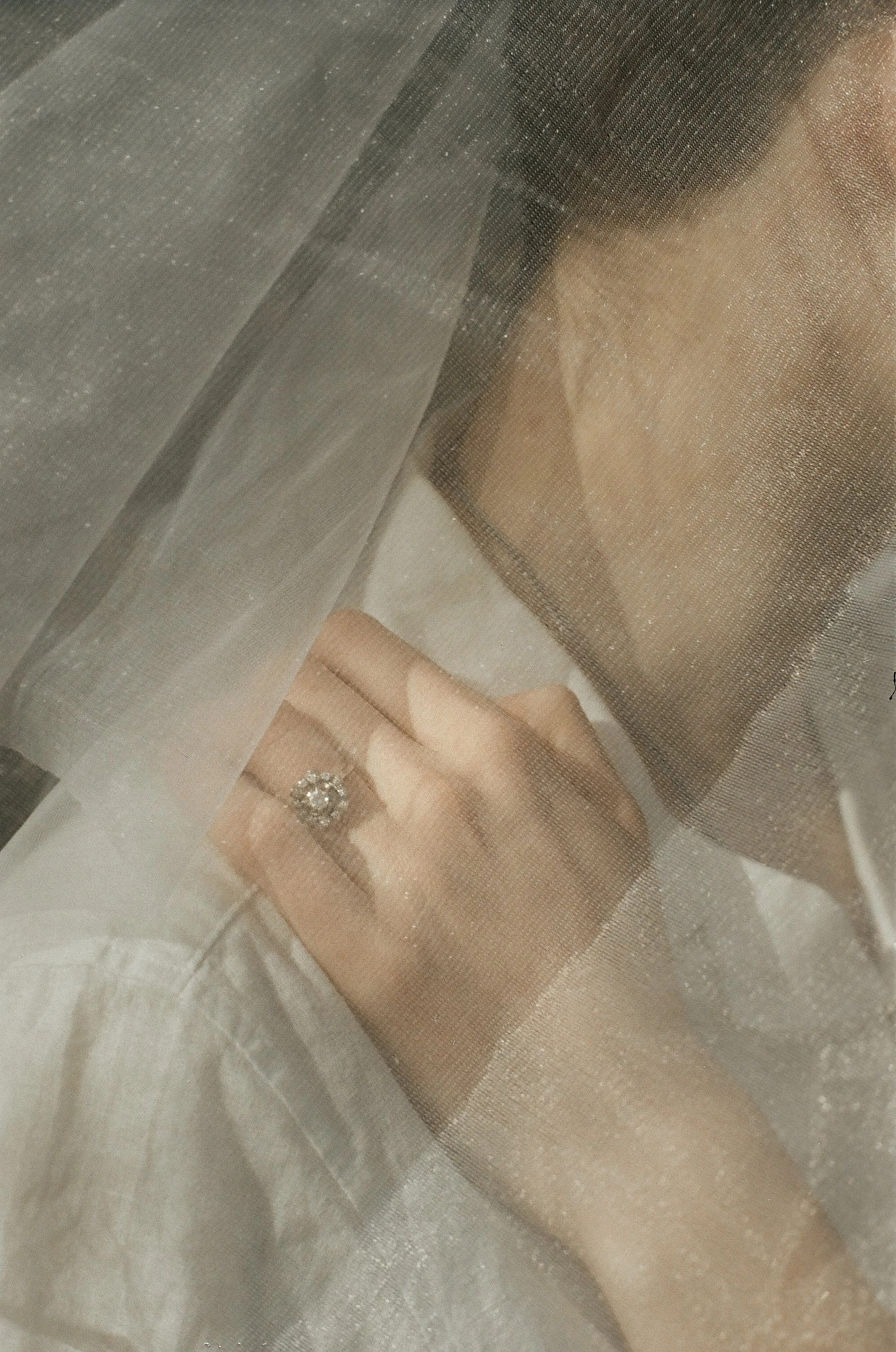 A person with a diamond ring on their finger, seen through a sheer curtain, with their hand resting on a box or jewelry display.
