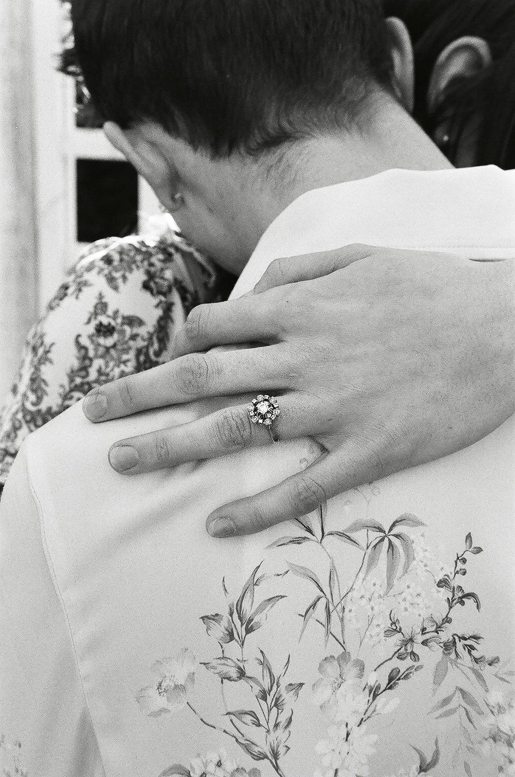 A black and white photo of a woman’s hand with an engagement ring resting on a man's shoulder. The woman’s face is not visible, and the man’s face is turned away. The background shows a bookcase and a floral-patterned garment.