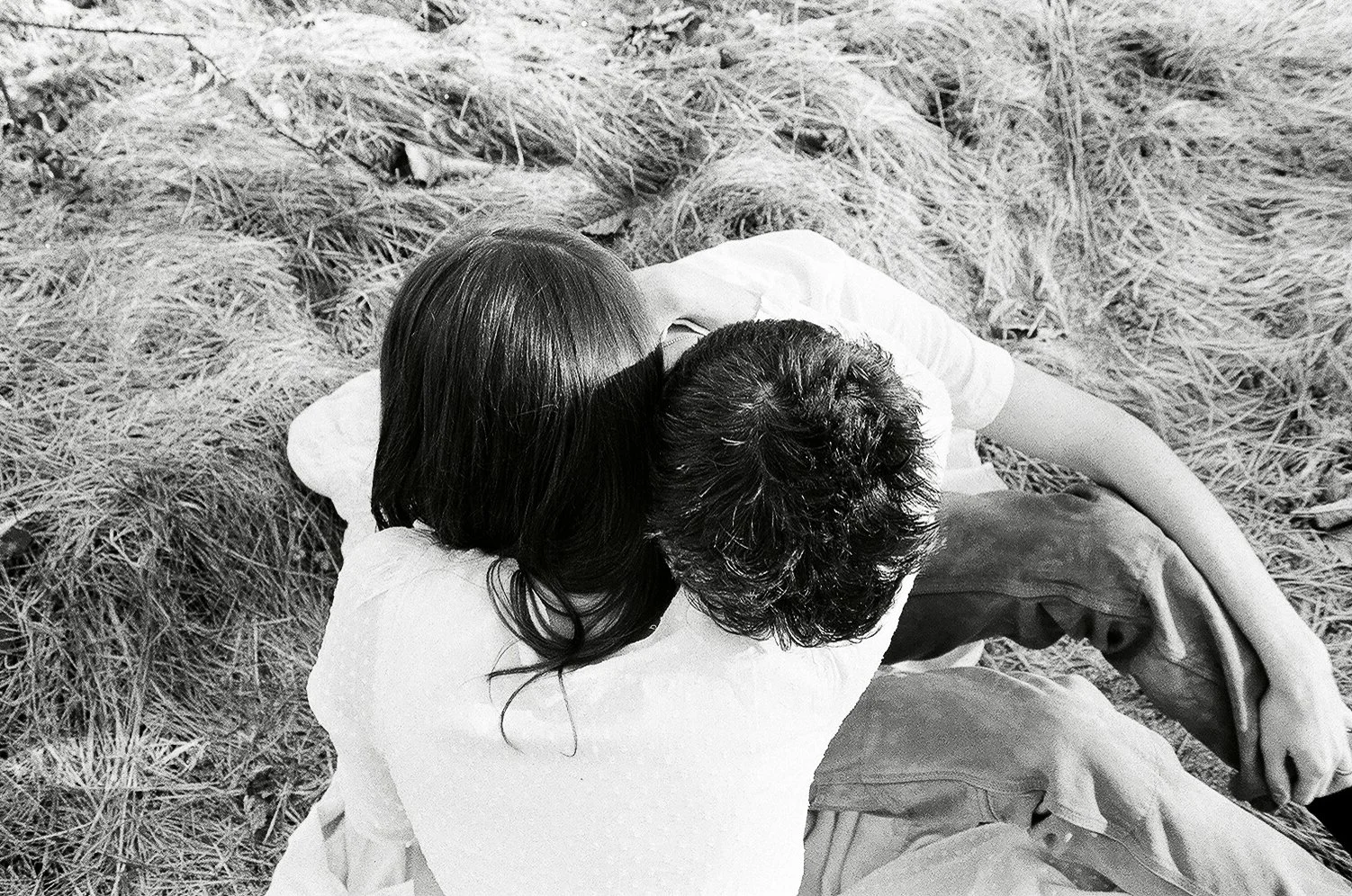 A black-and-white photo of two people hugging, sitting outdoors on dry grass.