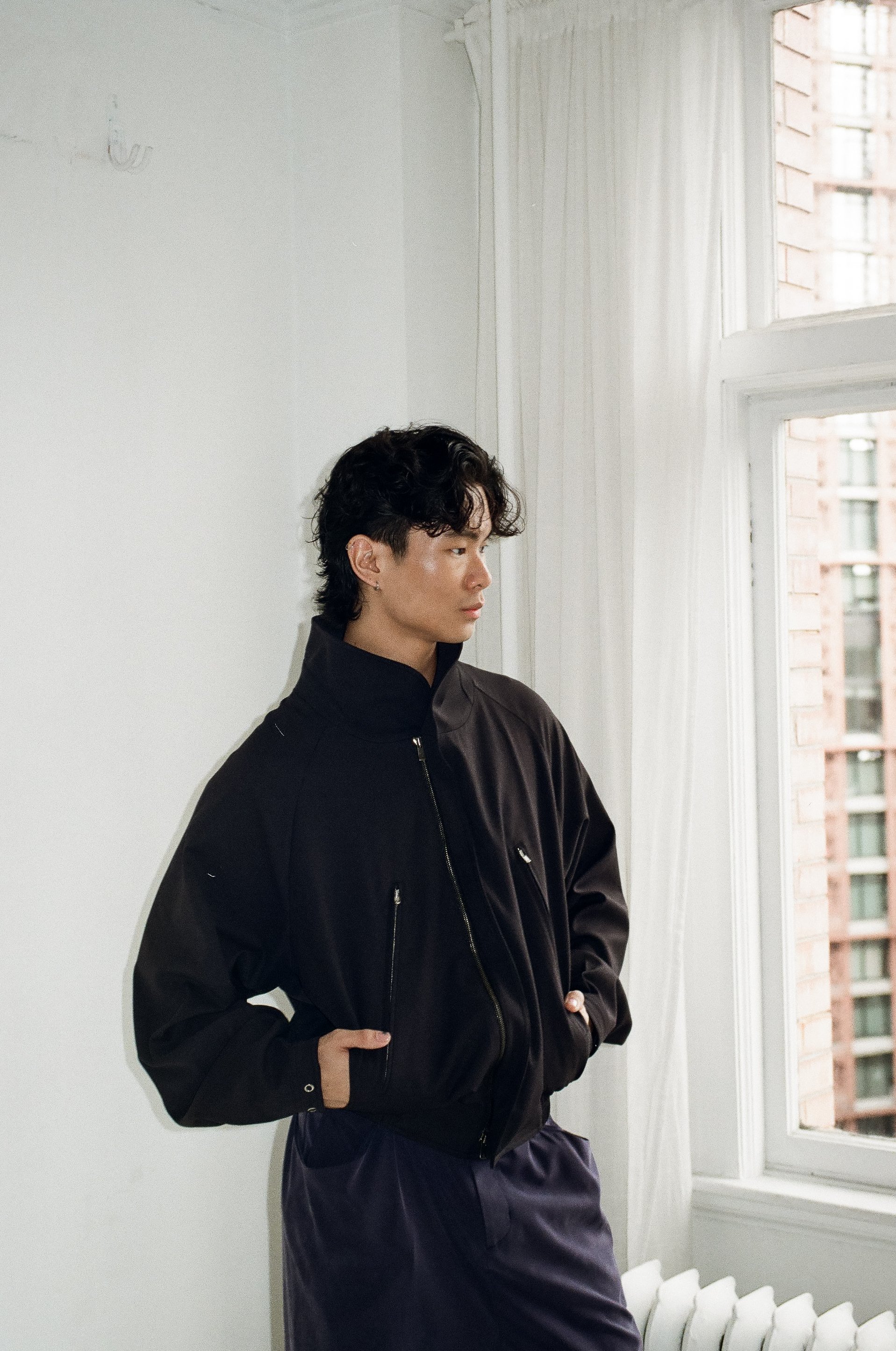 A young man with dark curly hair standing indoors by a large window with white curtains, wearing a black jacket with zipper details and dark pants.