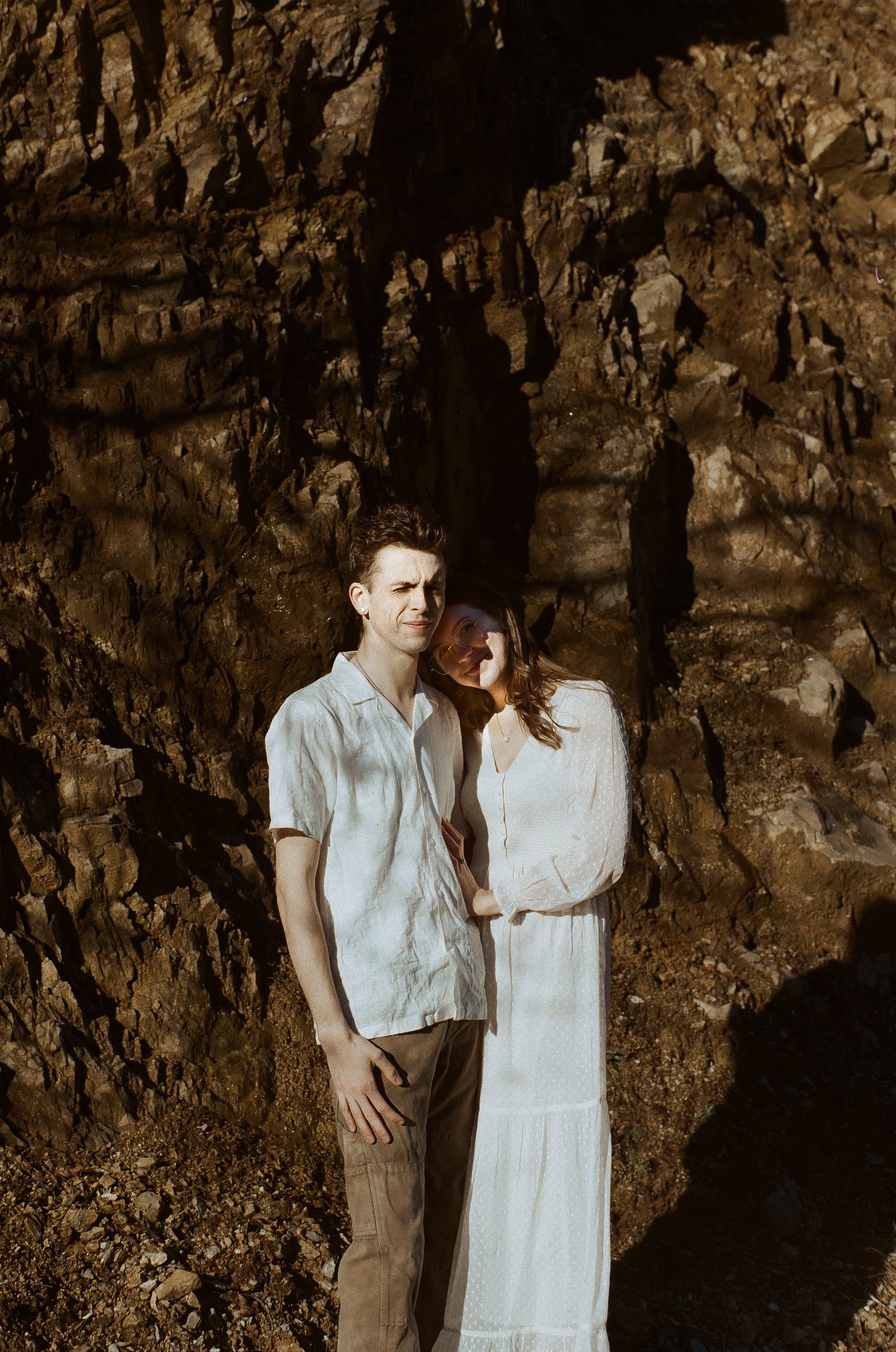 A young man and woman in light-colored clothing standing close together against a rocky, earth-toned natural backdrop with sunlight casting shadows.