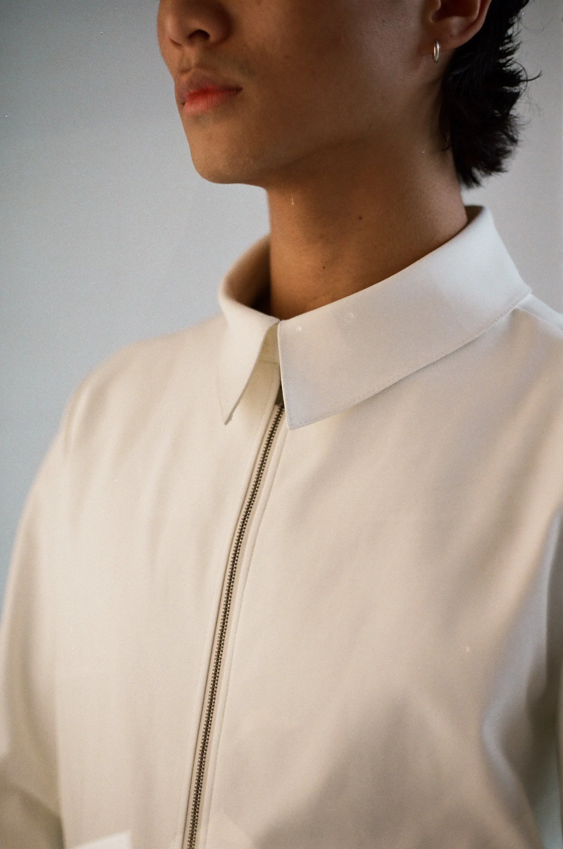 Close-up of a person wearing a white collared shirt with a zipper front, seen from the neck to just below the lips, with a small hoop earring visible on the right ear.