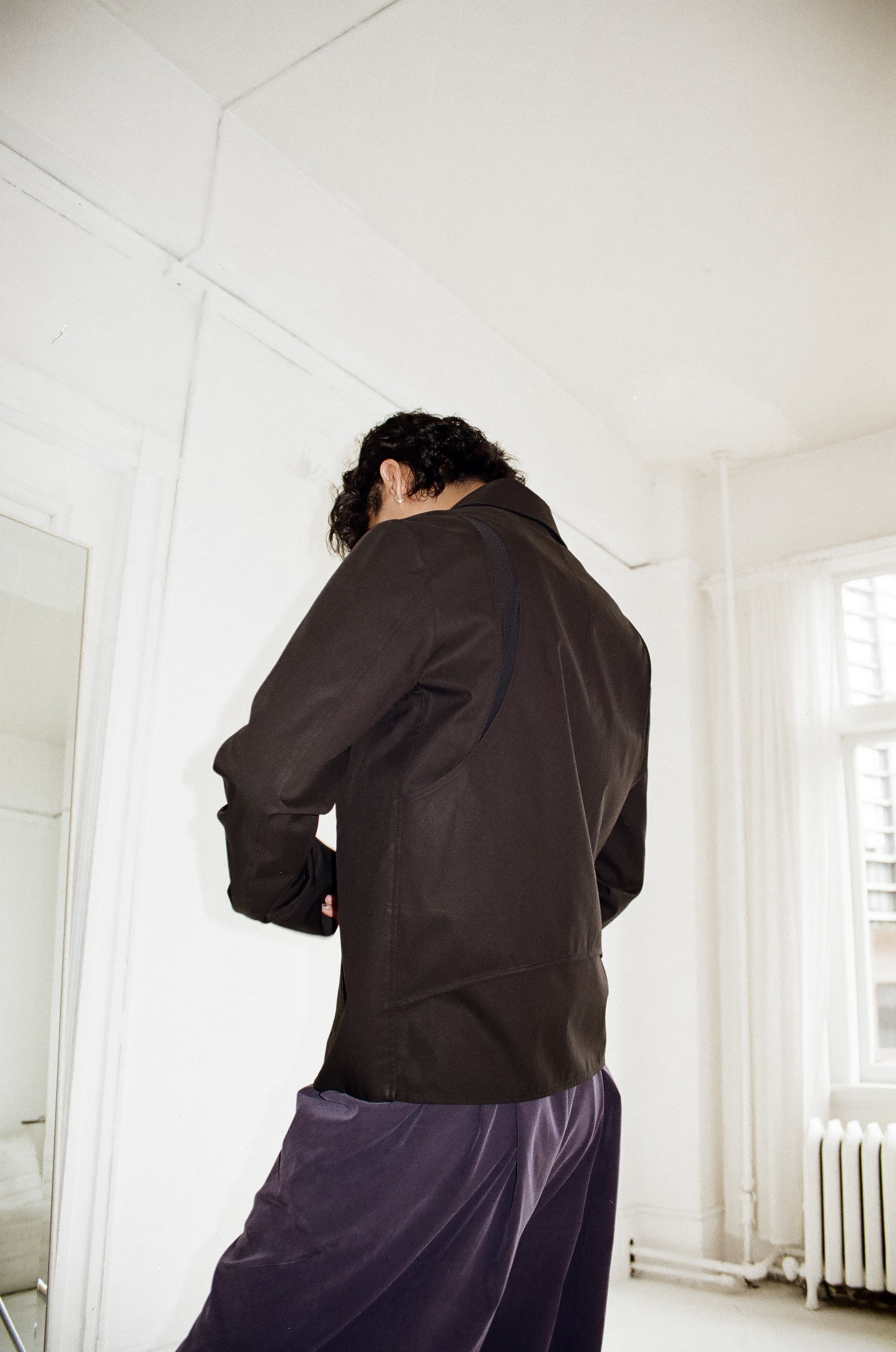 Back view of a person with curly hair wearing a dark brown jacket and purple pants, standing in a bright room with white walls, a window, and a radiator.