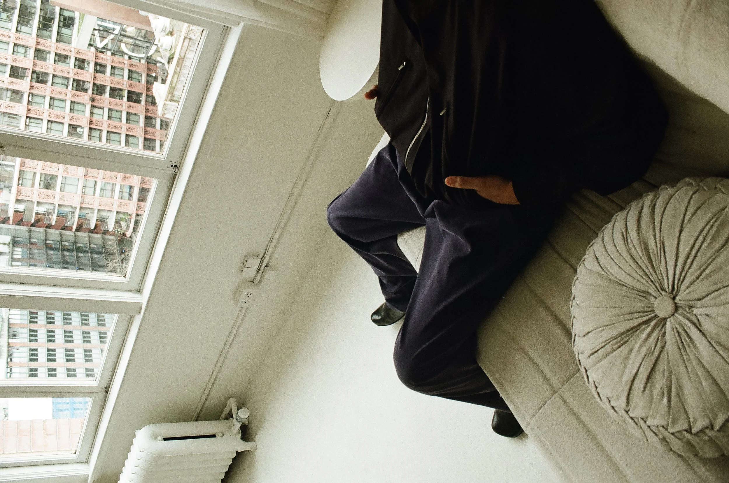 Two people sitting on a beige sofa near a large window, with a view of tall buildings outside, one person wearing dark pants and shoes, the other a beige skirt with a ruffled scrunch detail.