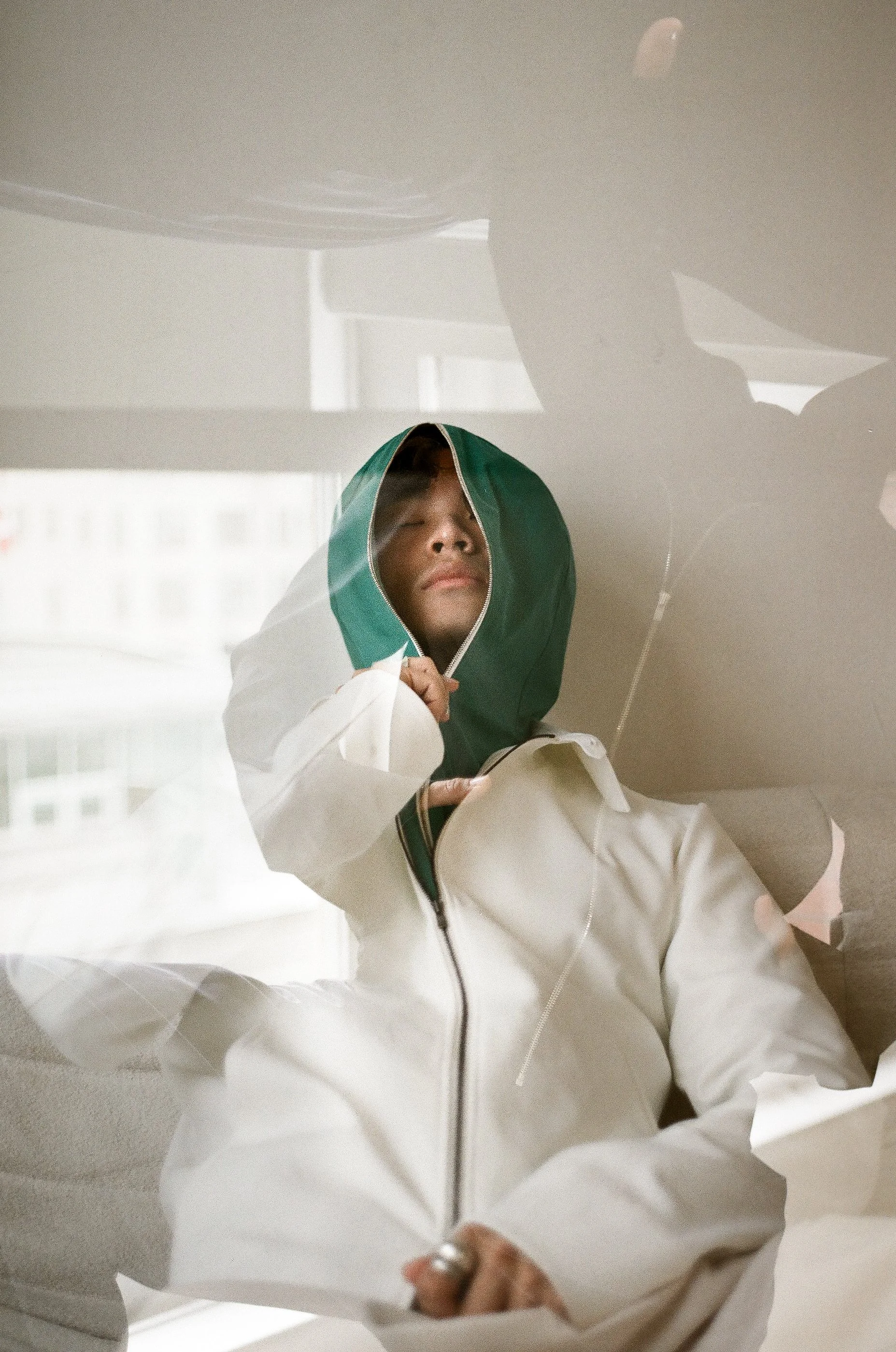 Person wearing a white jacket with a green hood, sitting with their eyes closed, reflected through a glass window.